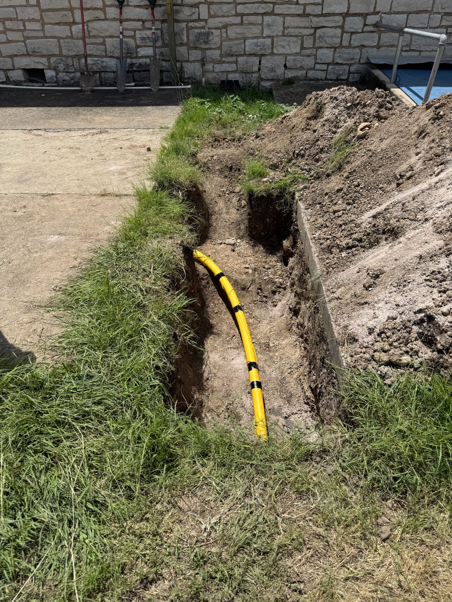 Yellow gas line in a narrow trench dug through grass and dirt, near a brick wall and concrete.