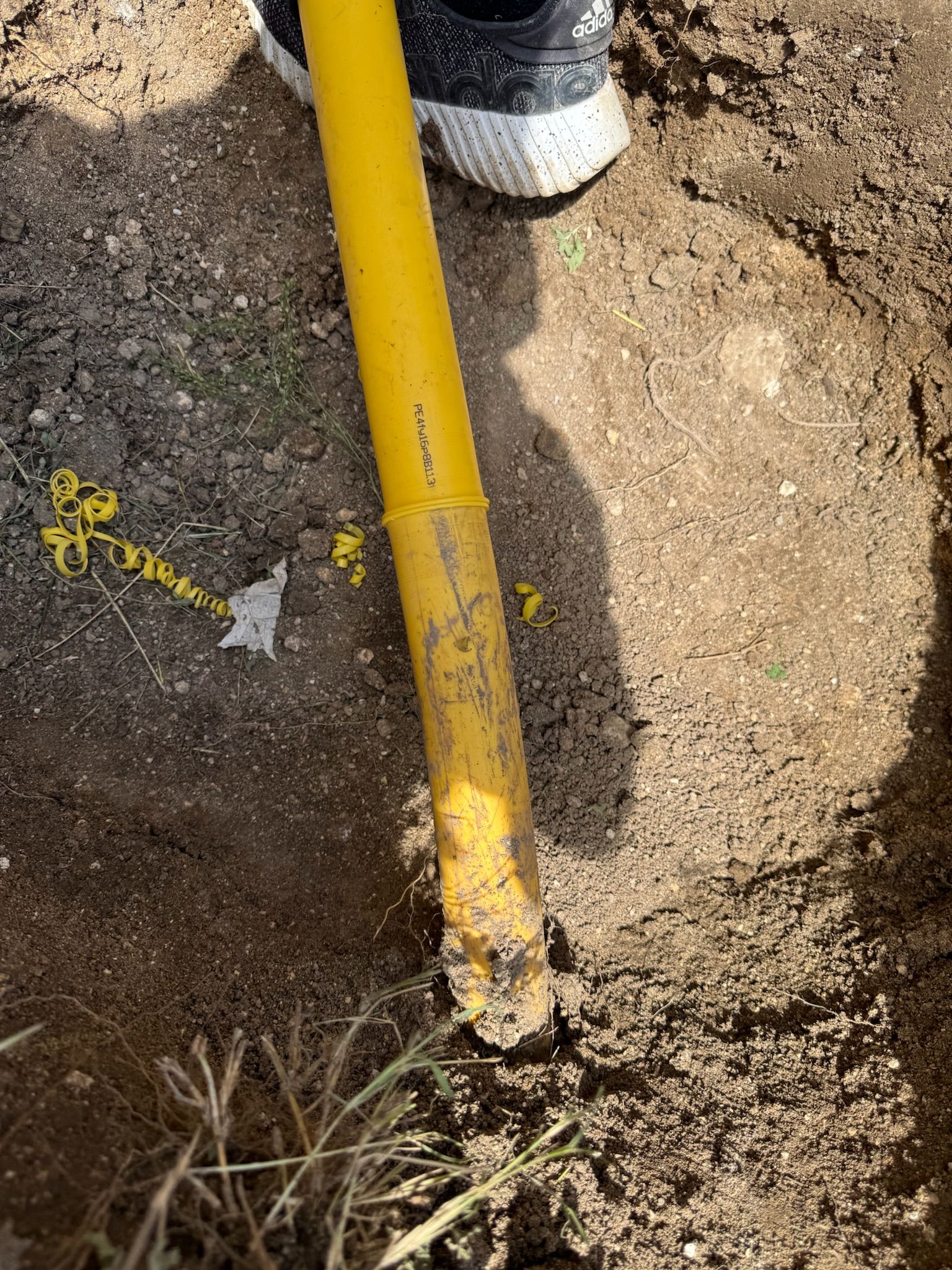 Yellow shovel in a hole dug in dirt, with a person's shoe in the upper corner.