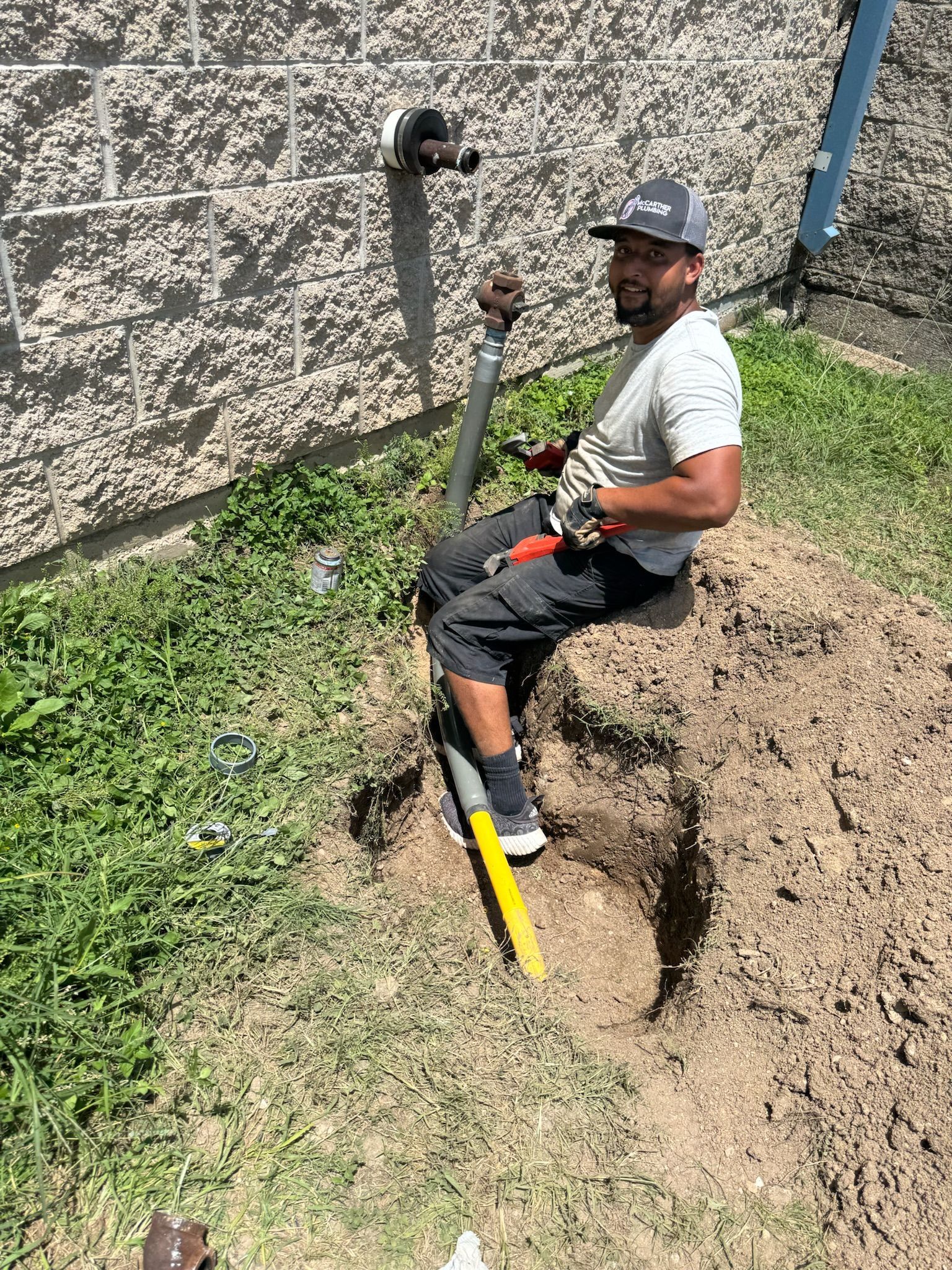 Man digging in a yard near a building, wearing work gloves and a cap, with a shovel.