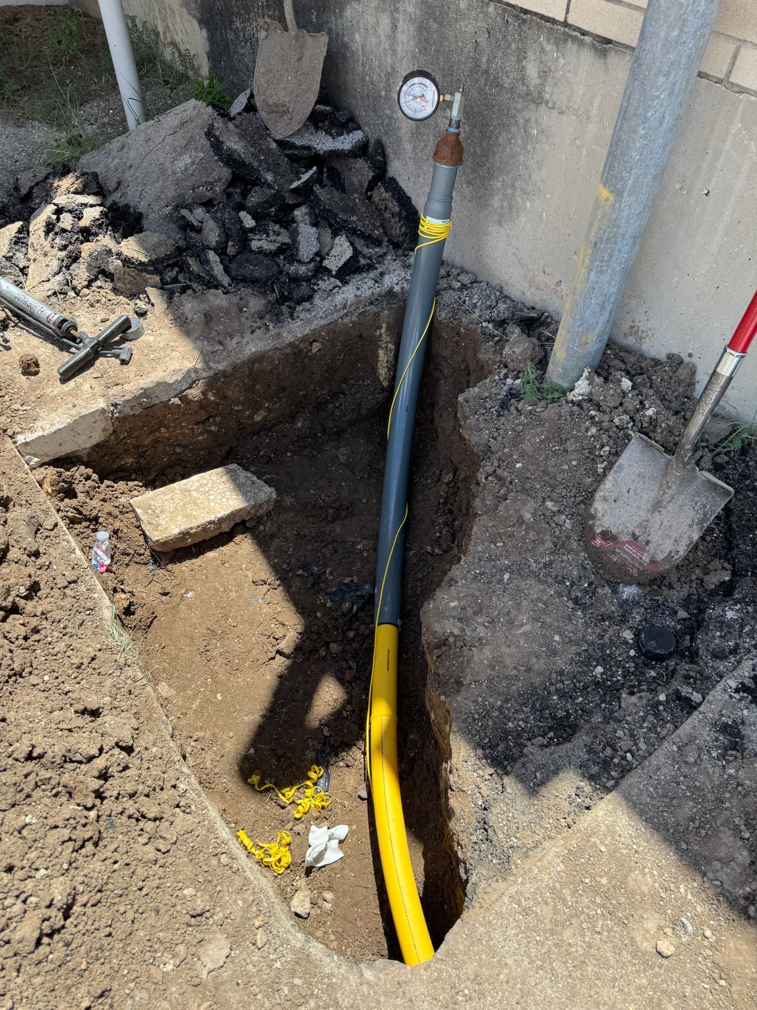 A trench dug in the ground with a yellow and black gas line pipe running through it and a gauge attached.