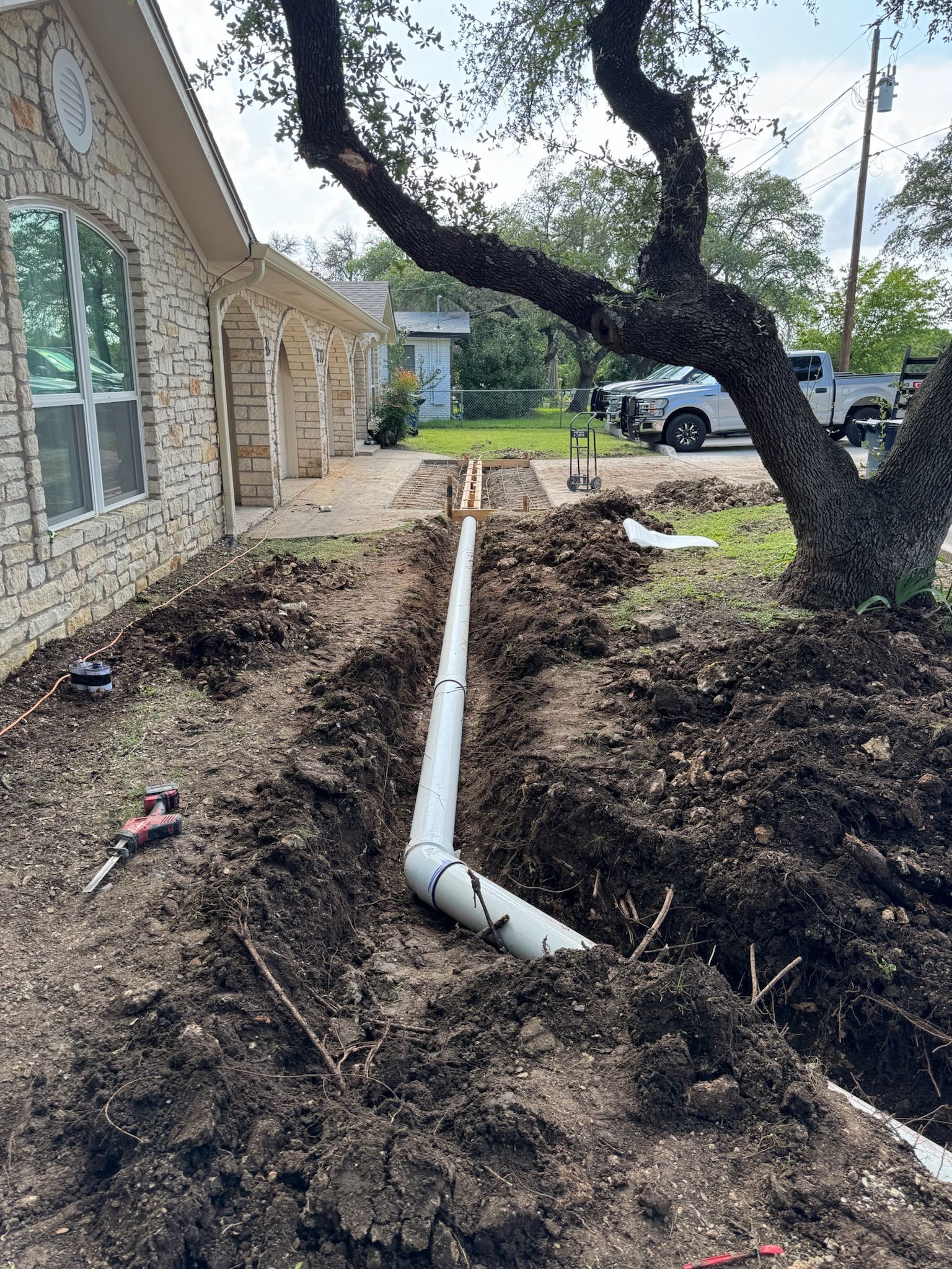Trench dug along house with white pipe installed; lawn, tree, and vehicle in background.