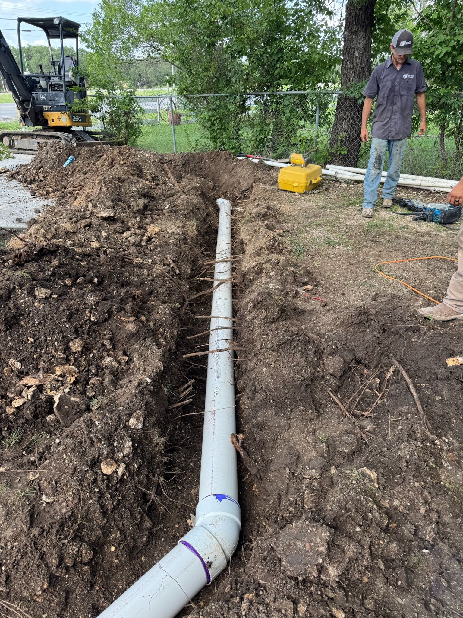 A trench with white pipe, and a worker is standing nearby. A small excavator is visible.