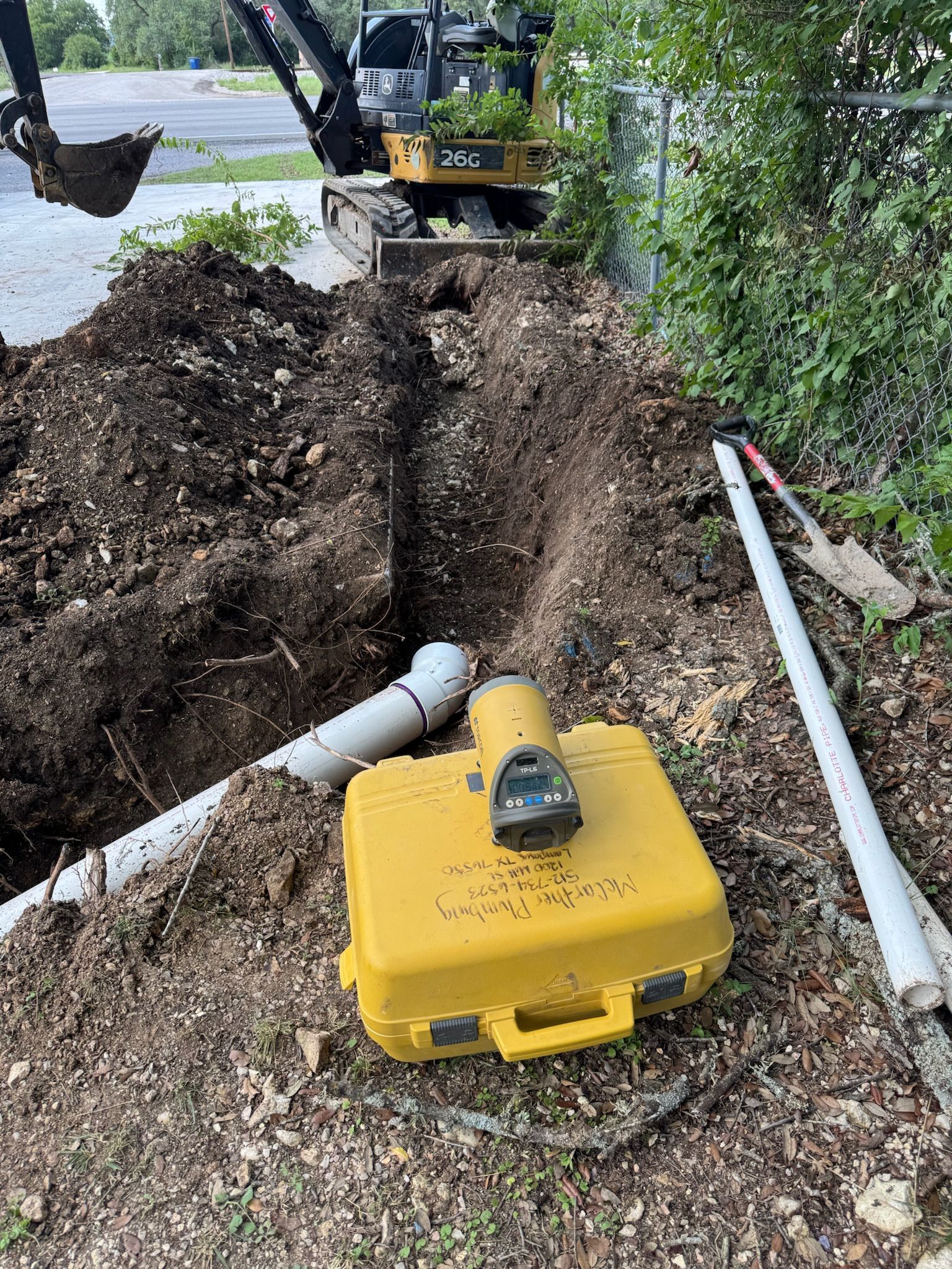 Trench with white pipes, yellow level case, and small excavator. Outdoors, daytime.