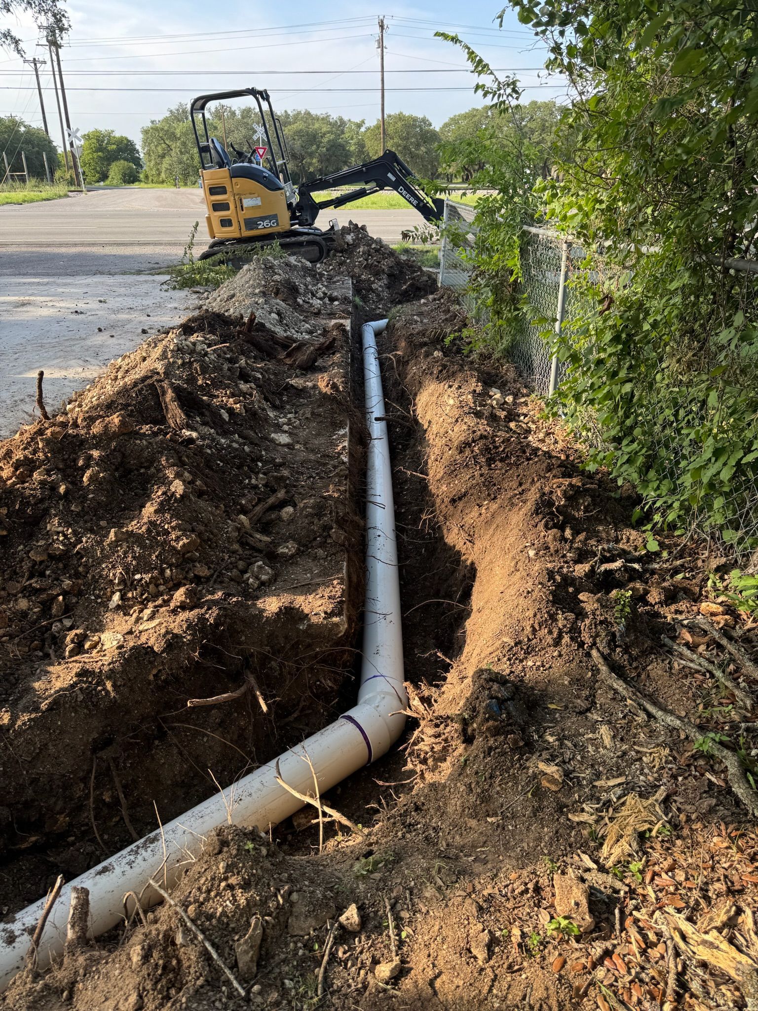 Trench with drainage pipe being installed; excavator in the background, dirt and vegetation surround the scene.