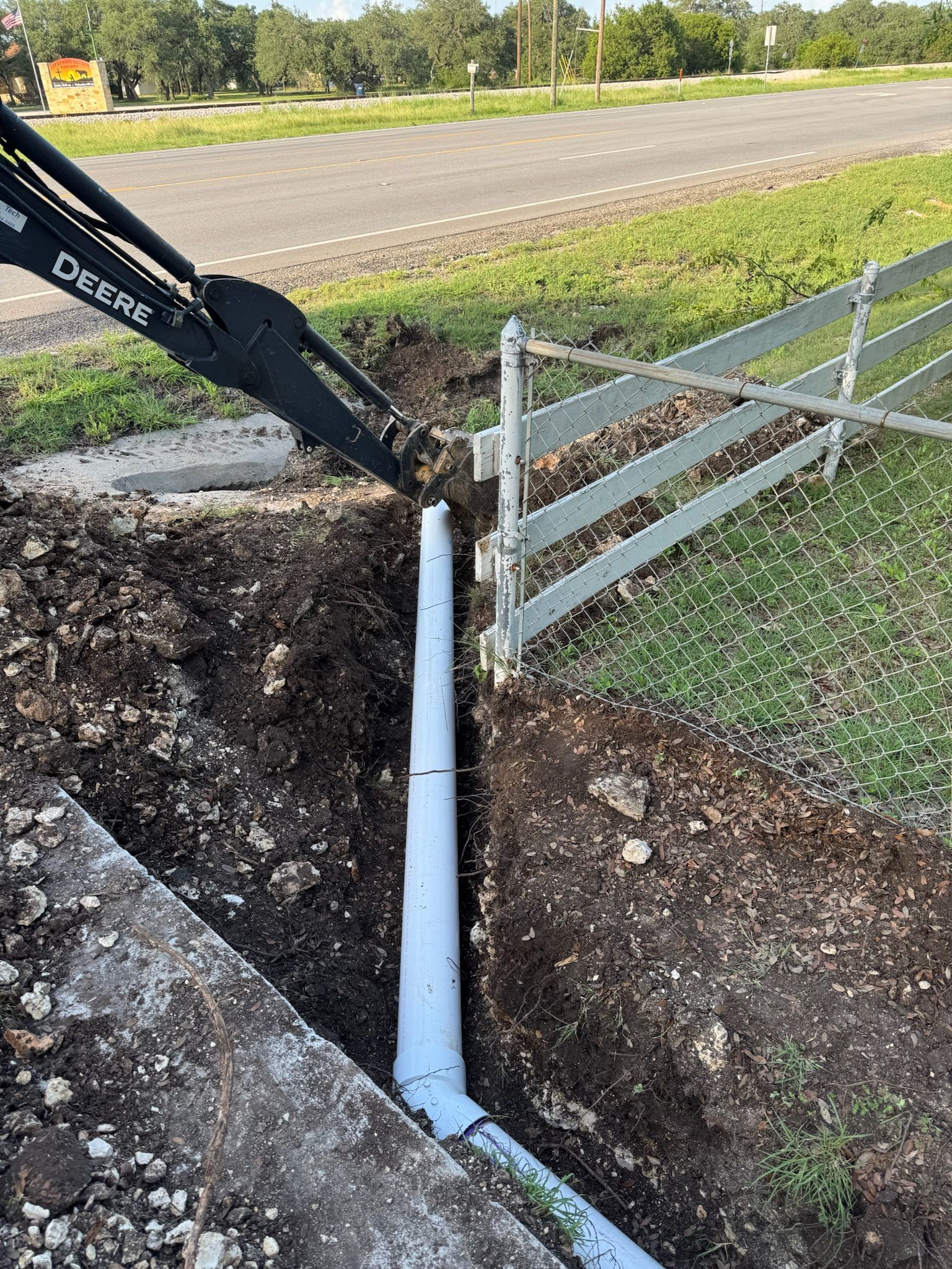A backhoe installing drainage pipe beside a fence and road.