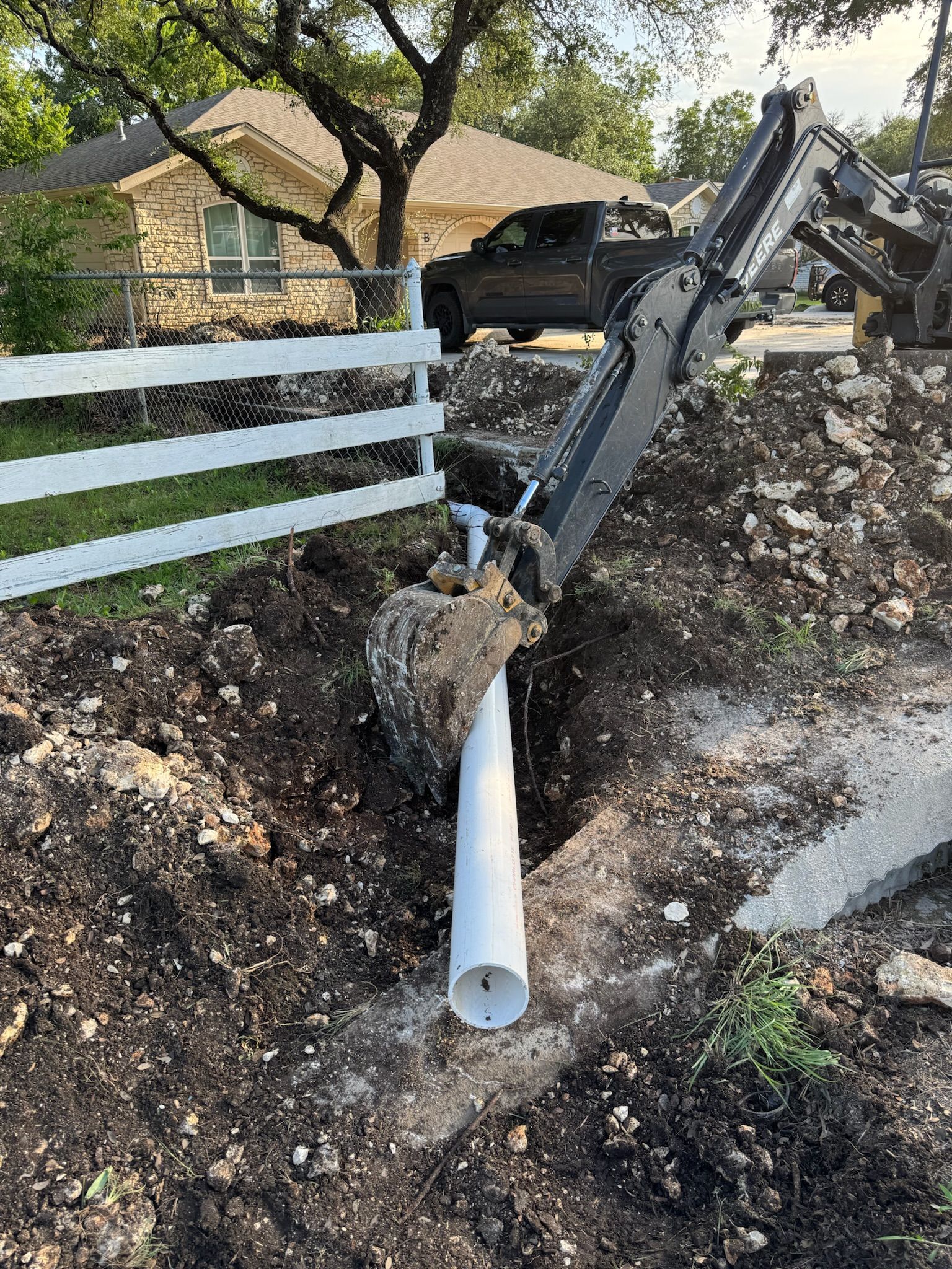 An excavator placing a white pipe into a trench in a yard, with a house and fence in the background.