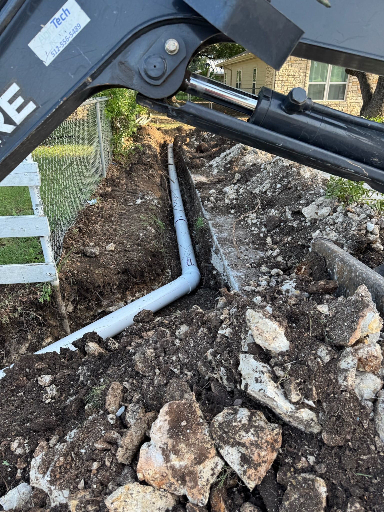 A tractor installing a perforated drain pipe in a trench next to a fence.