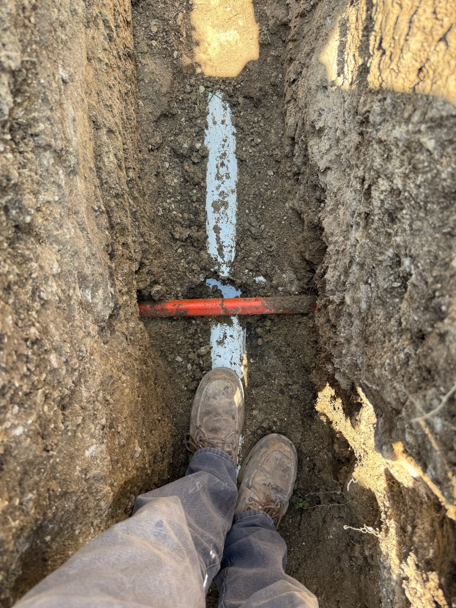 A trench with a white pipe, red pipe, and muddy boots.