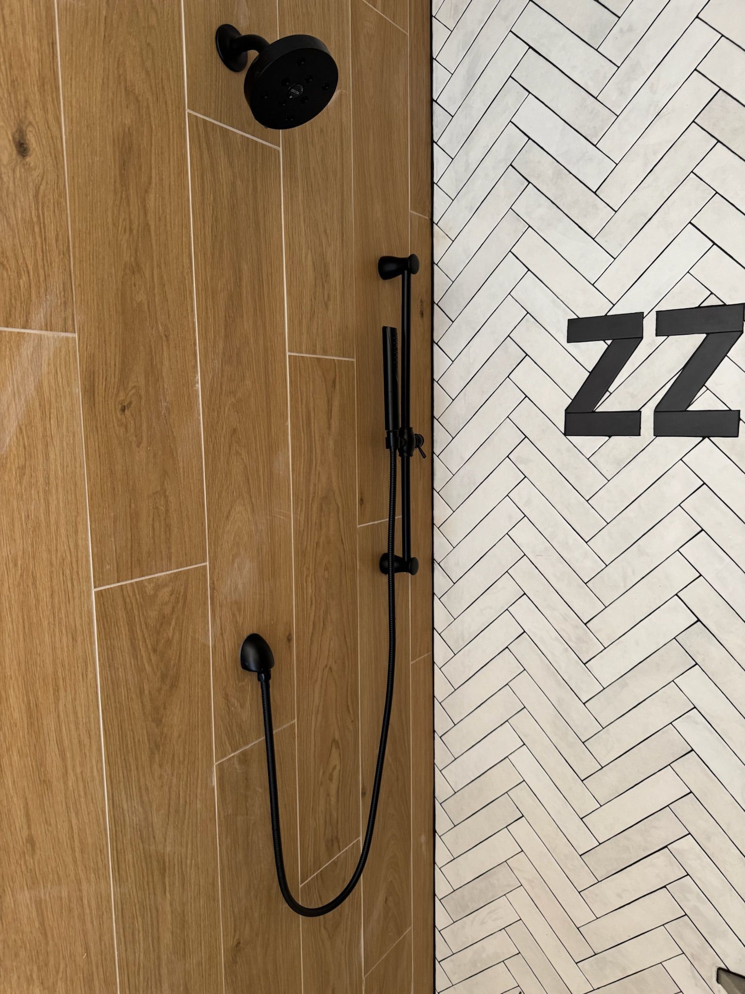 Black shower head and wand on wood-look tile next to a herringbone-tiled wall.