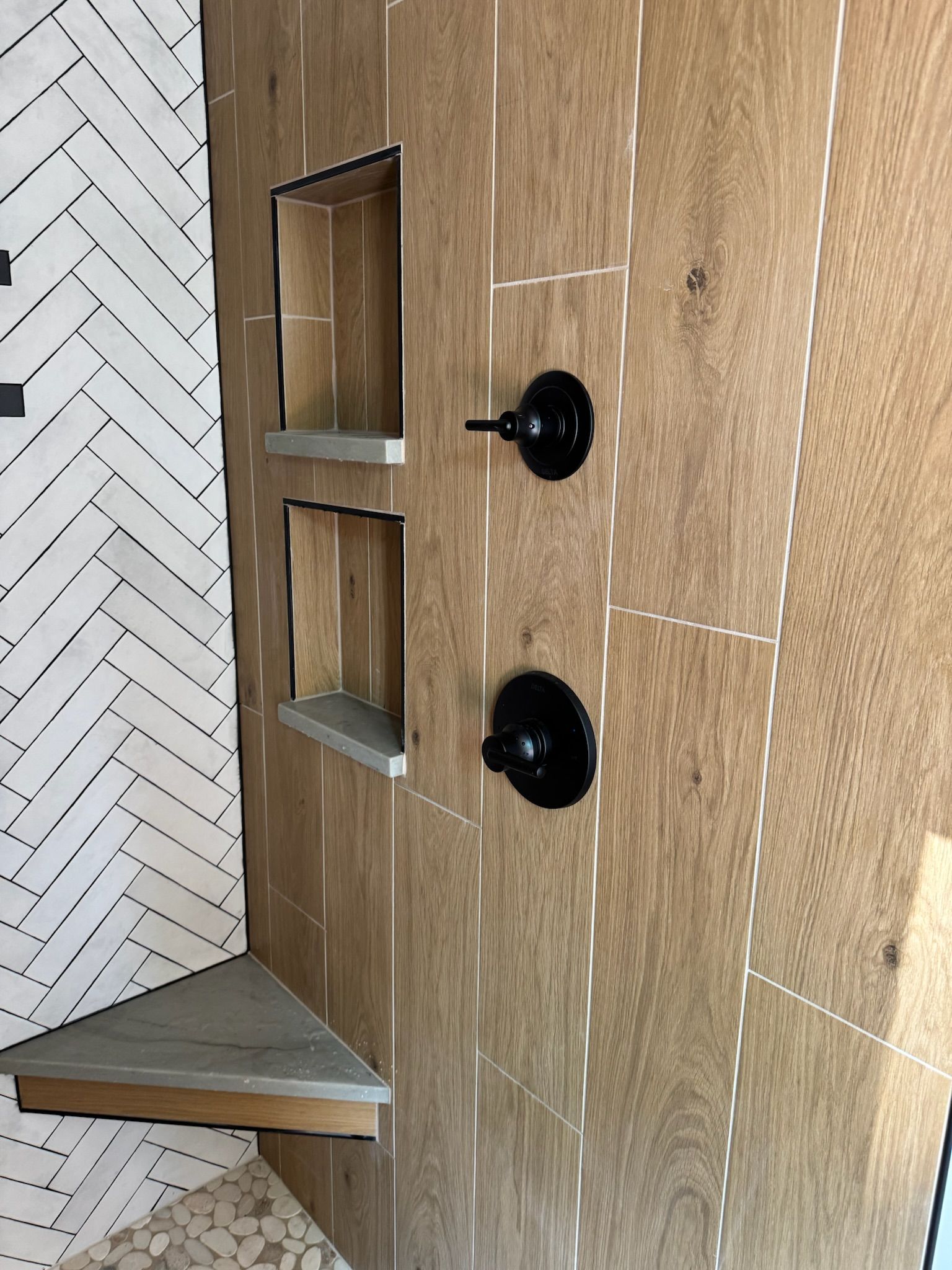 Shower with wood-look tile, built-in shelves, black fixtures, and herringbone pattern accent wall.