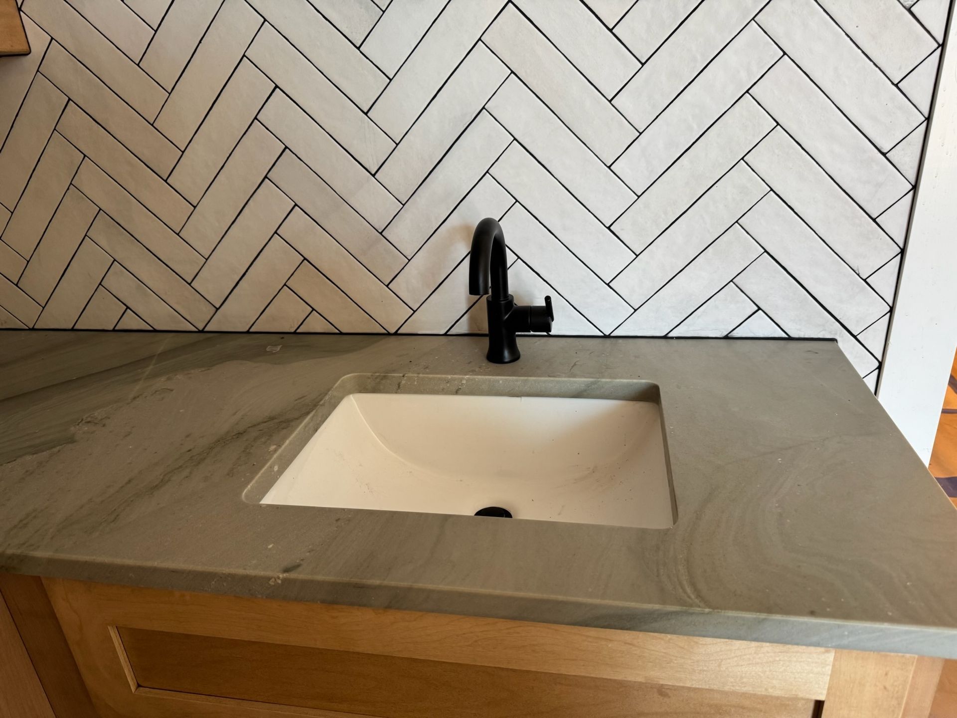 Bathroom vanity with a white rectangular sink, black faucet, gray countertop, and herringbone tile backsplash.