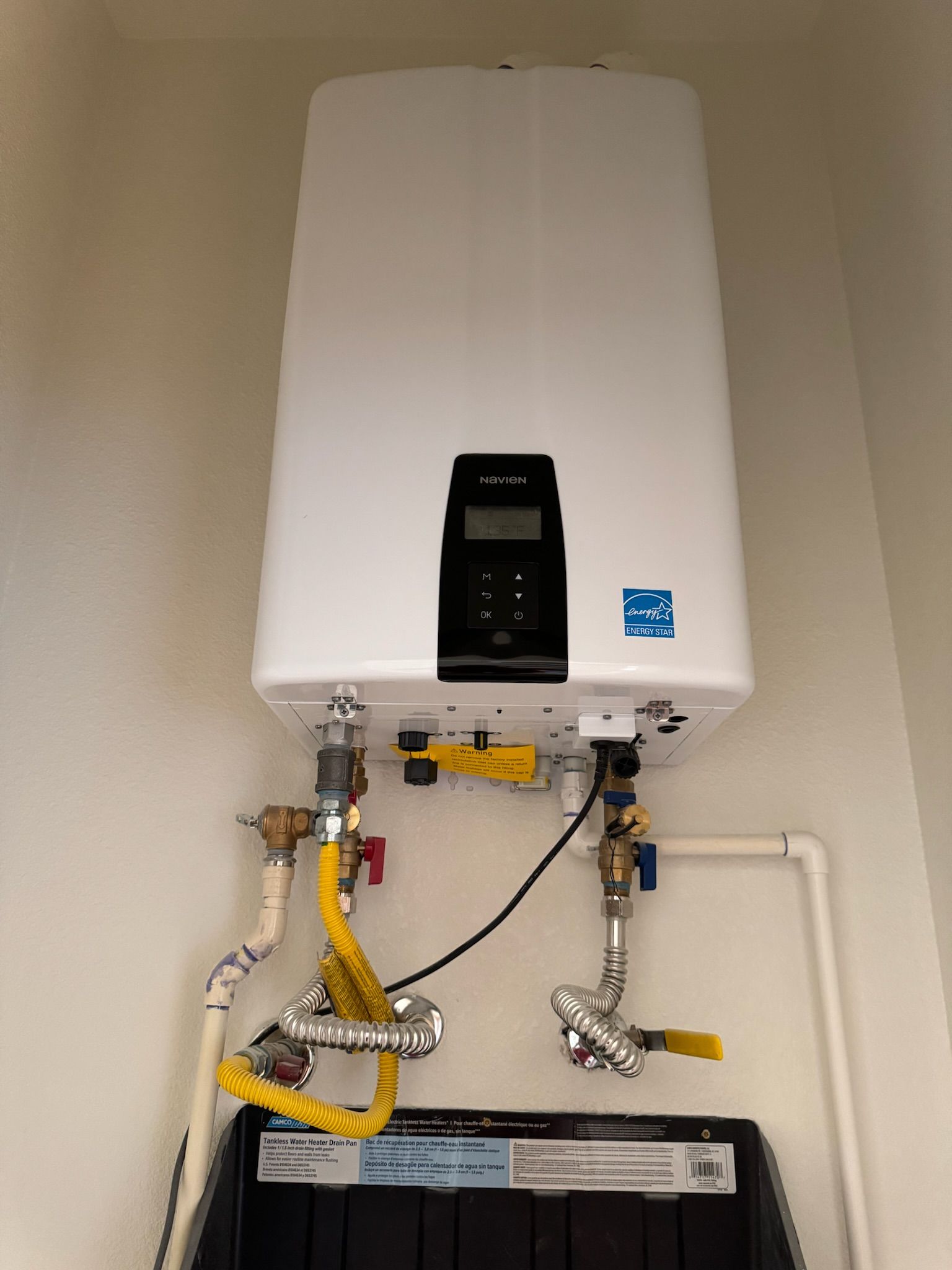 White tankless water heater mounted on a wall with connected pipes and control panel.