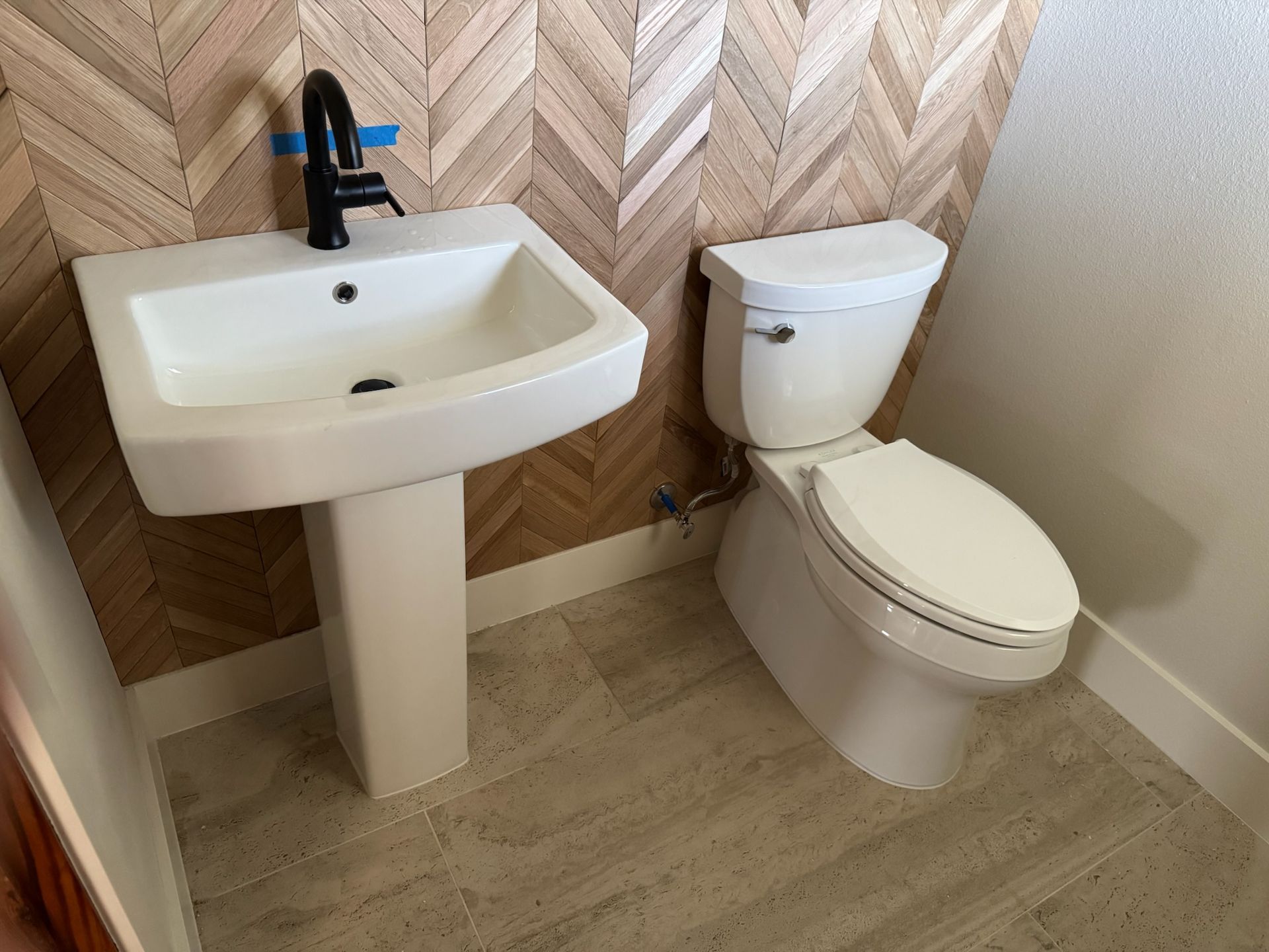 A small bathroom with a pedestal sink, black faucet, and toilet. Herringbone wood-look wall.