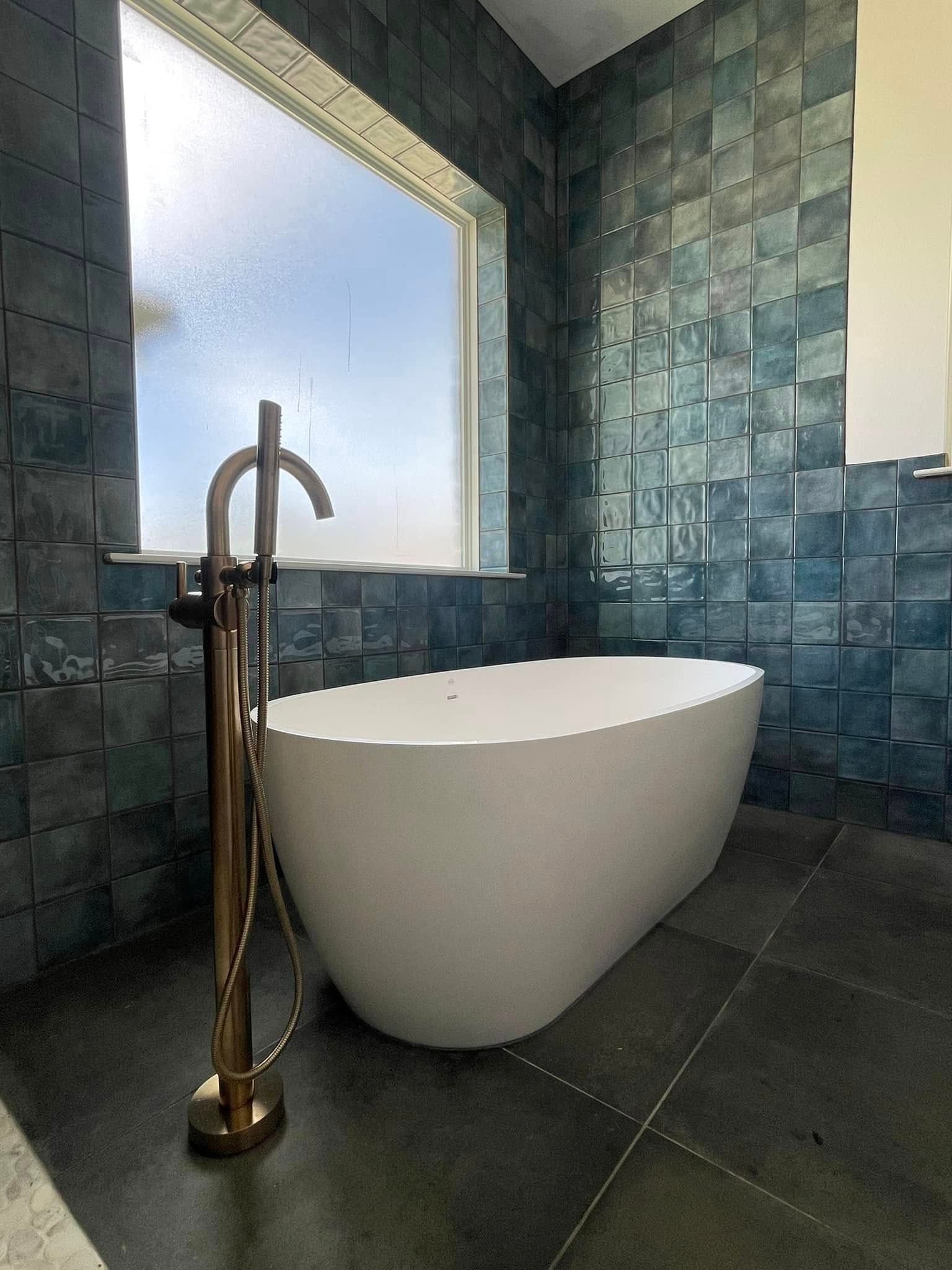 Freestanding white bathtub in a bathroom with blue tiled walls, gold faucet, and window.