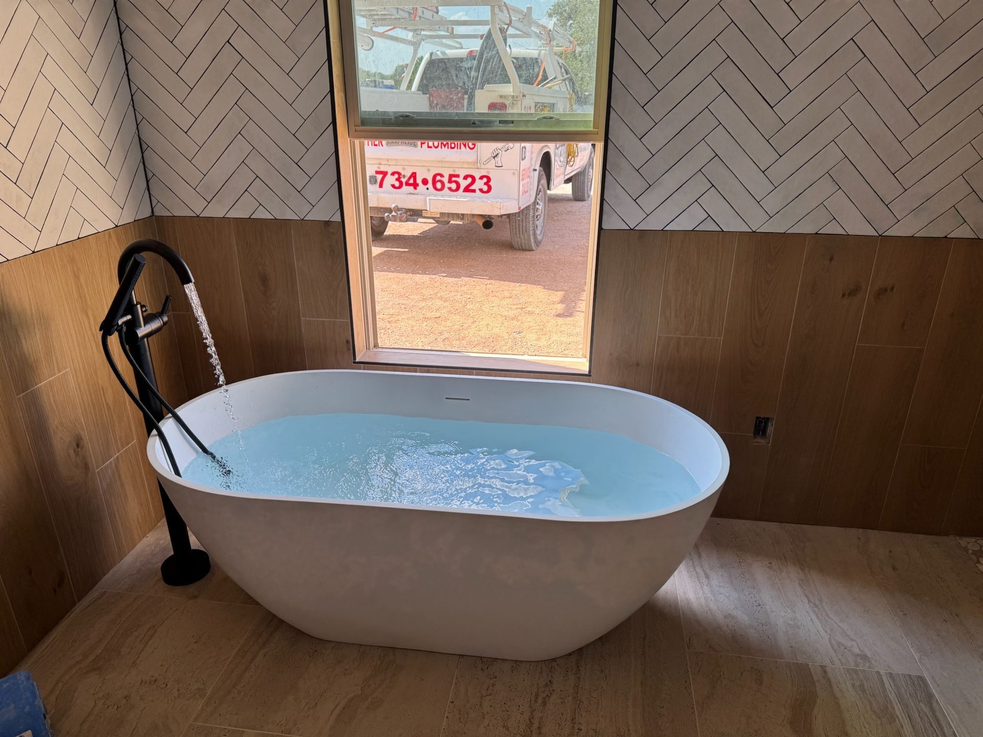 Freestanding white bathtub filled with water, next to an open window, with patterned wall tile.