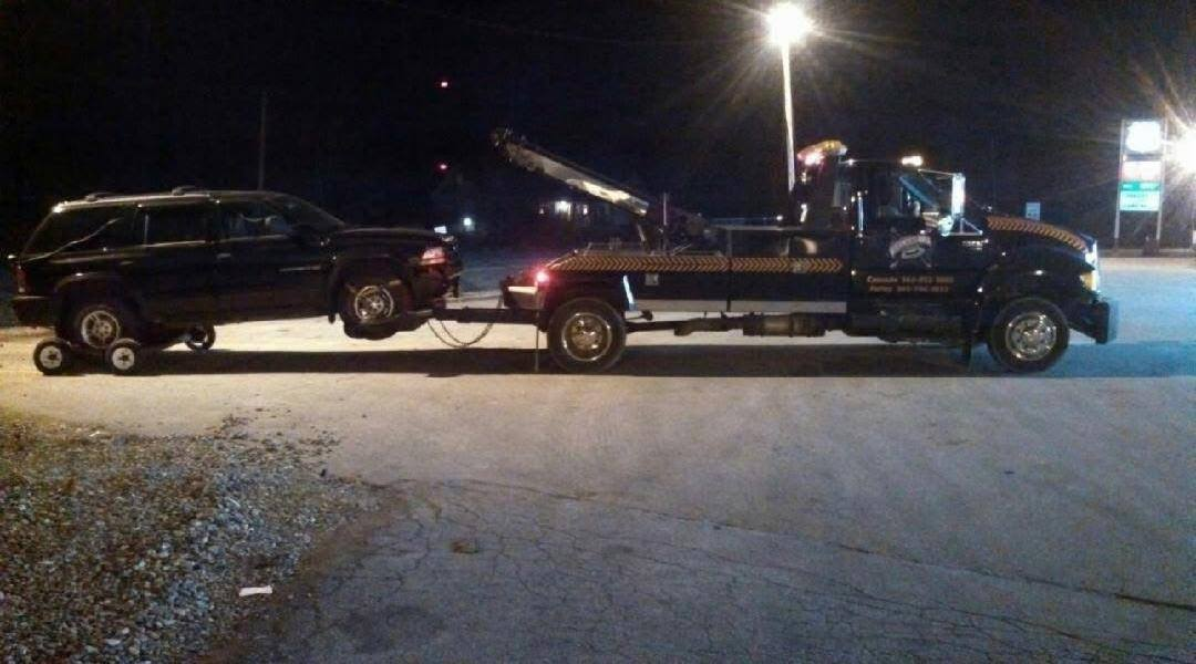 A black suv is being towed by a tow truck at night.