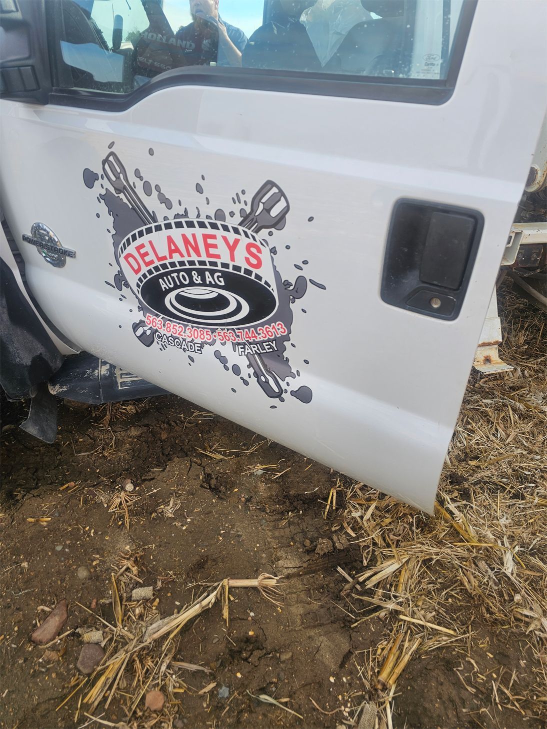 White truck door with 'Delaney's Auto & Ag' logo, set on dirt and stubble.
