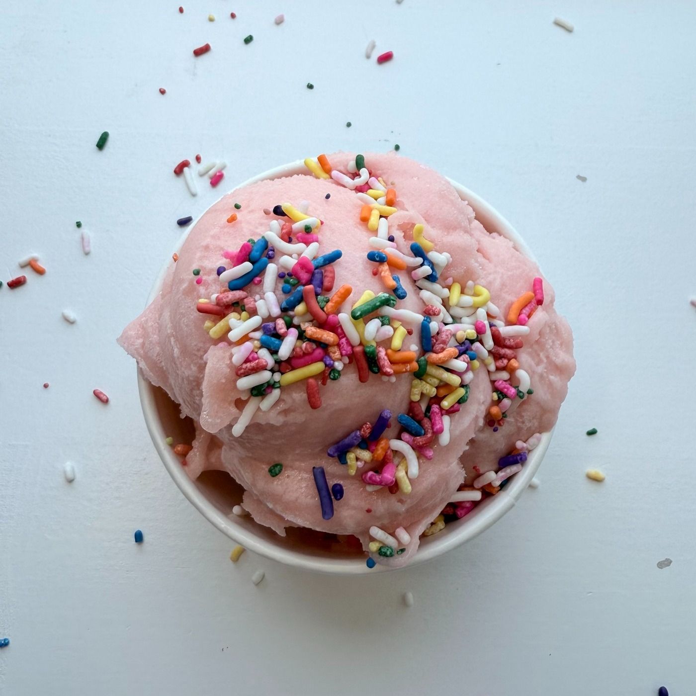 Pink ice cream with sprinkles in a white bowl on a white surface.