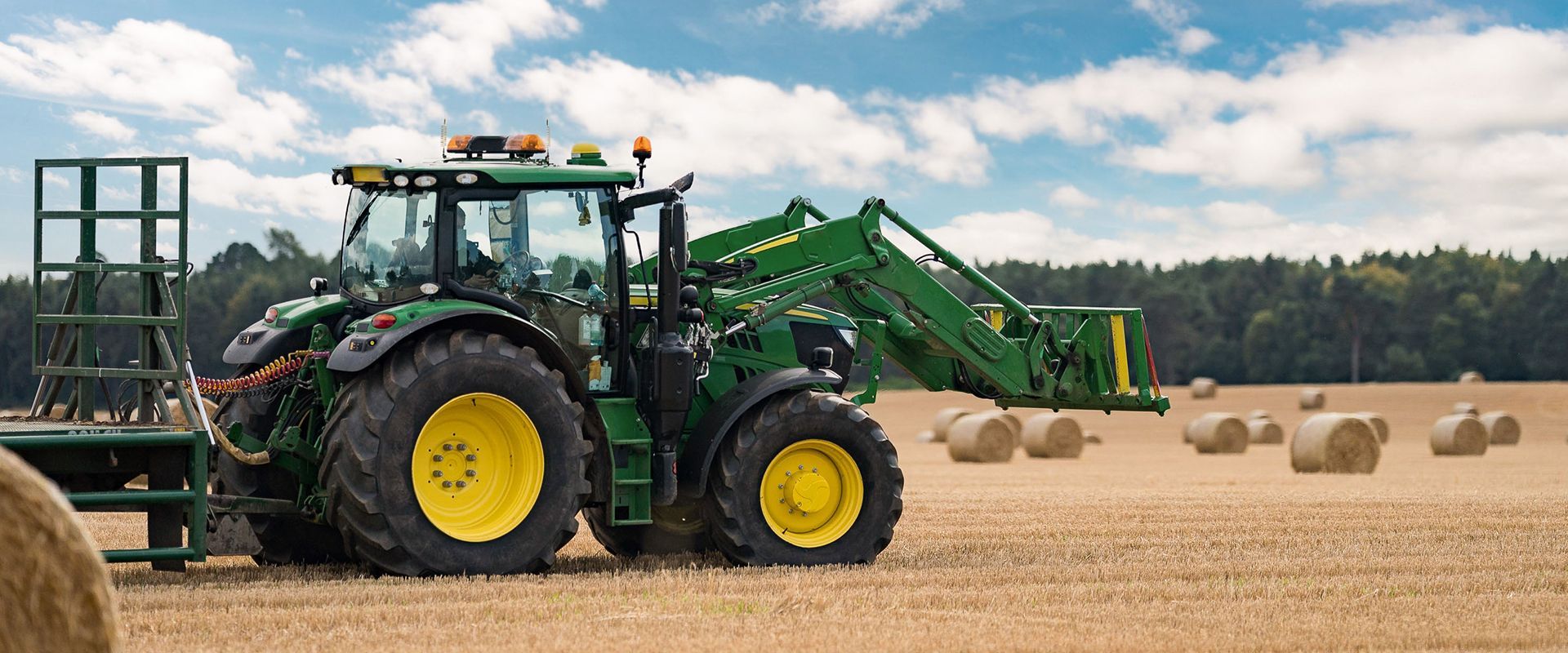 A green tractor is driving through a field of hay bales.