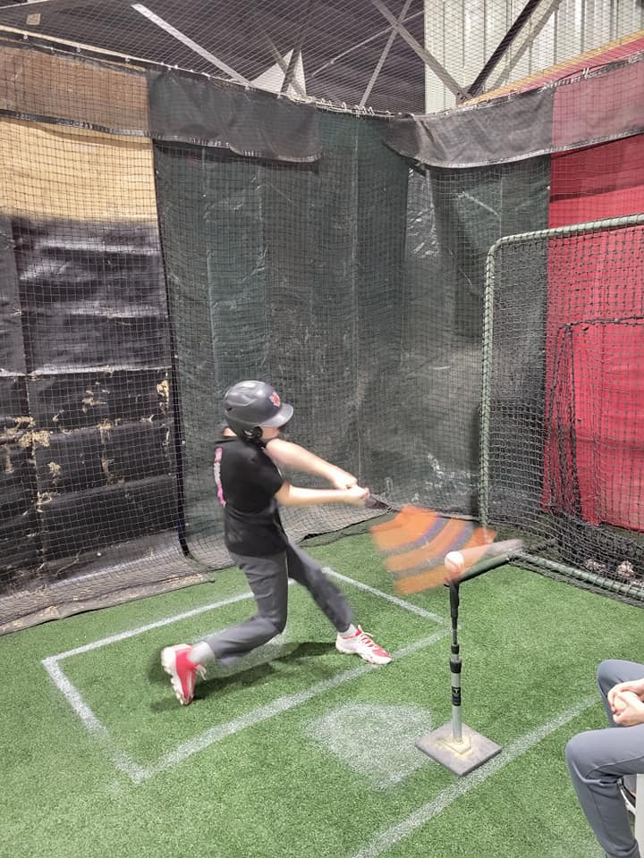 A young girl is swinging a bat at a baseball in a batting cage.
