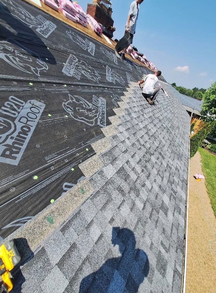 Roofers installing shingles on a house roof. One worker on the roof, others near the chimney.