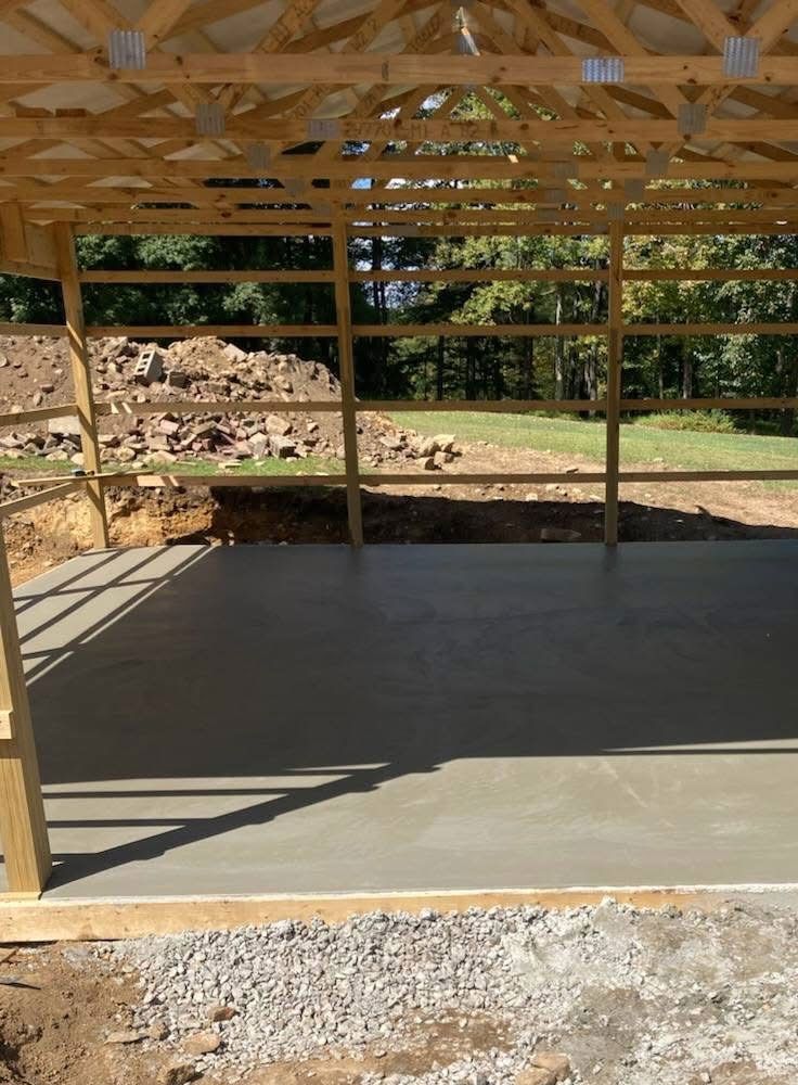 New concrete floor inside a wooden framed open-air structure, overlooking a grassy yard and woods.