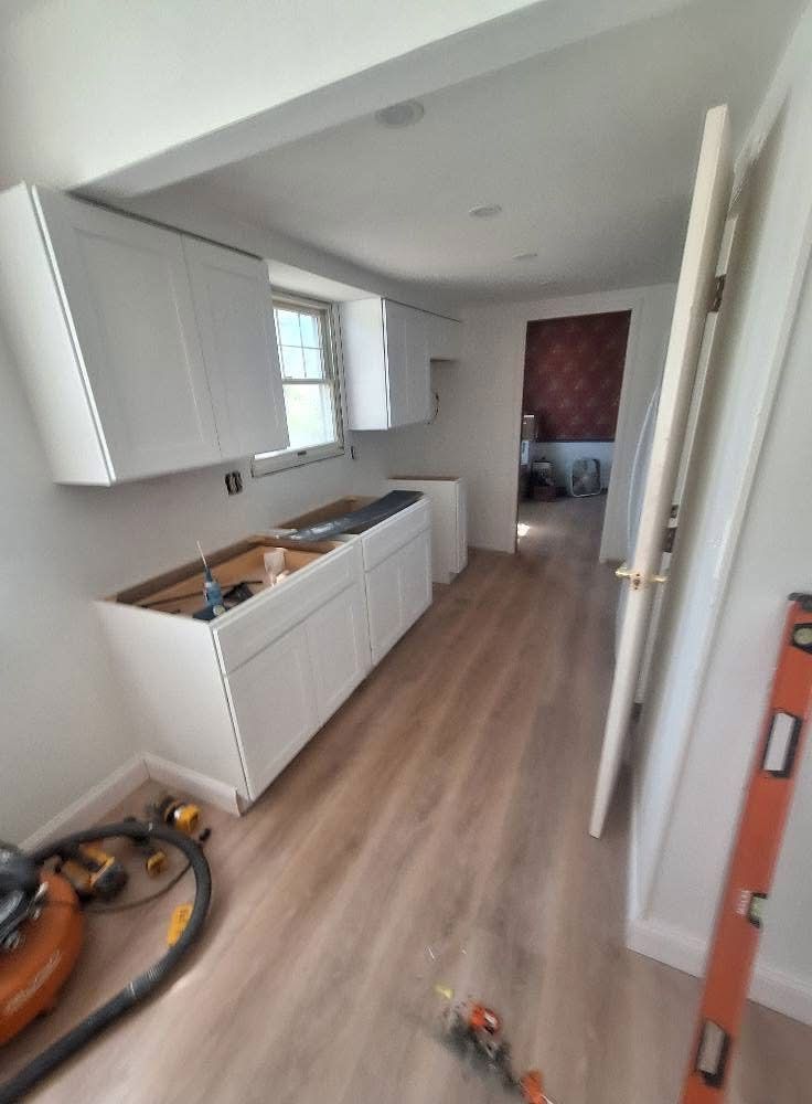 Kitchen remodel in progress: white cabinets, bare countertops, light wood-look flooring. Doorway leads to a hallway.