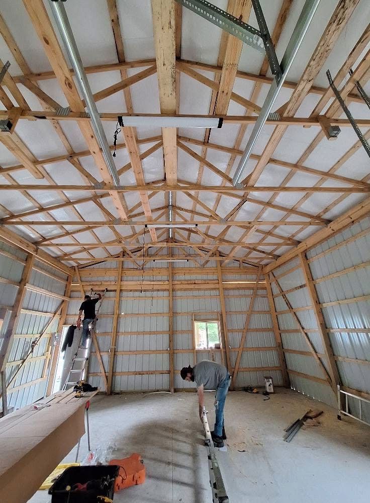 Two people work inside a large, unfinished wooden-framed building with metal siding and a high ceiling.