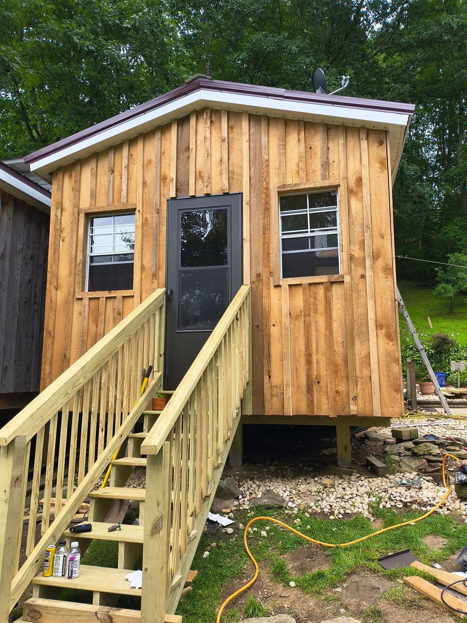 Small wooden cabin with stairs and a dark door, windows, and brown metal roof.