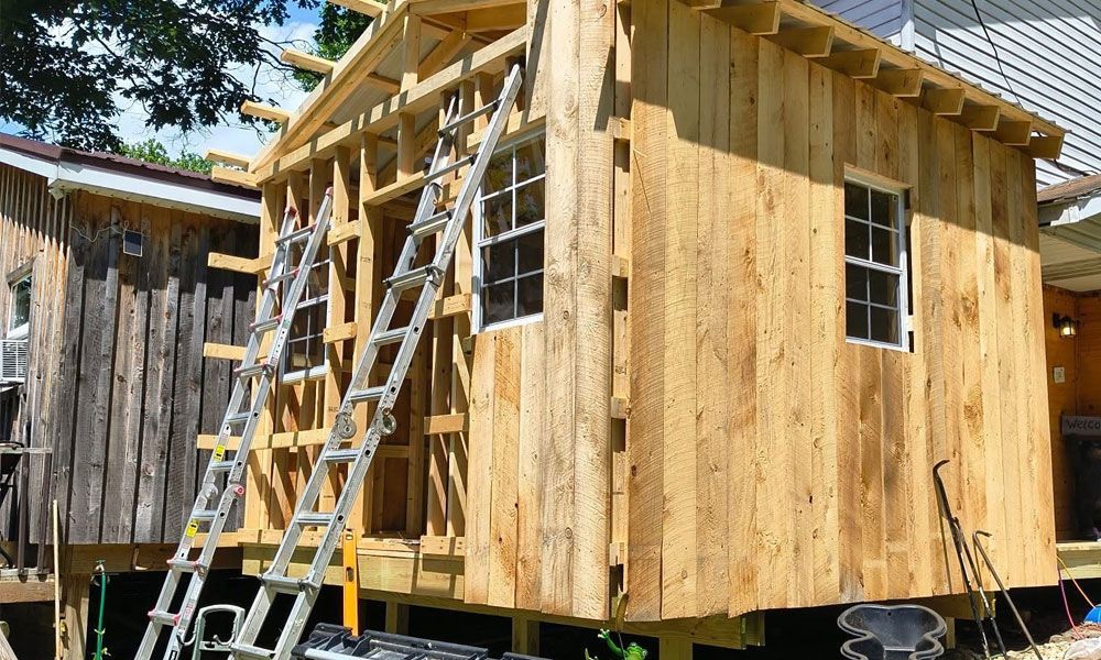 Wooden shed under construction, with ladders leaning against the frame, boards nailed to the exterior.
