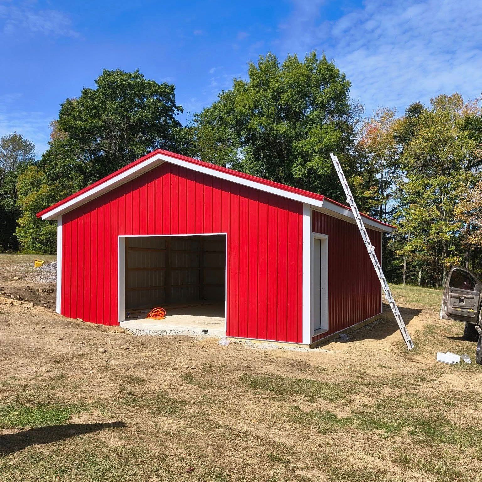 Red barn with open garage door, white trim, and a ladder, set in a field.