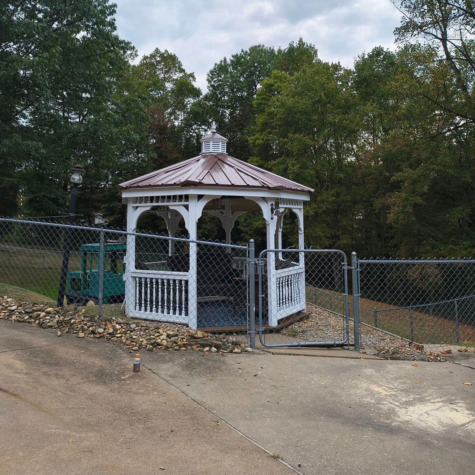 White gazebo with a copper roof, set back from a chain-link fence, surrounded by trees.