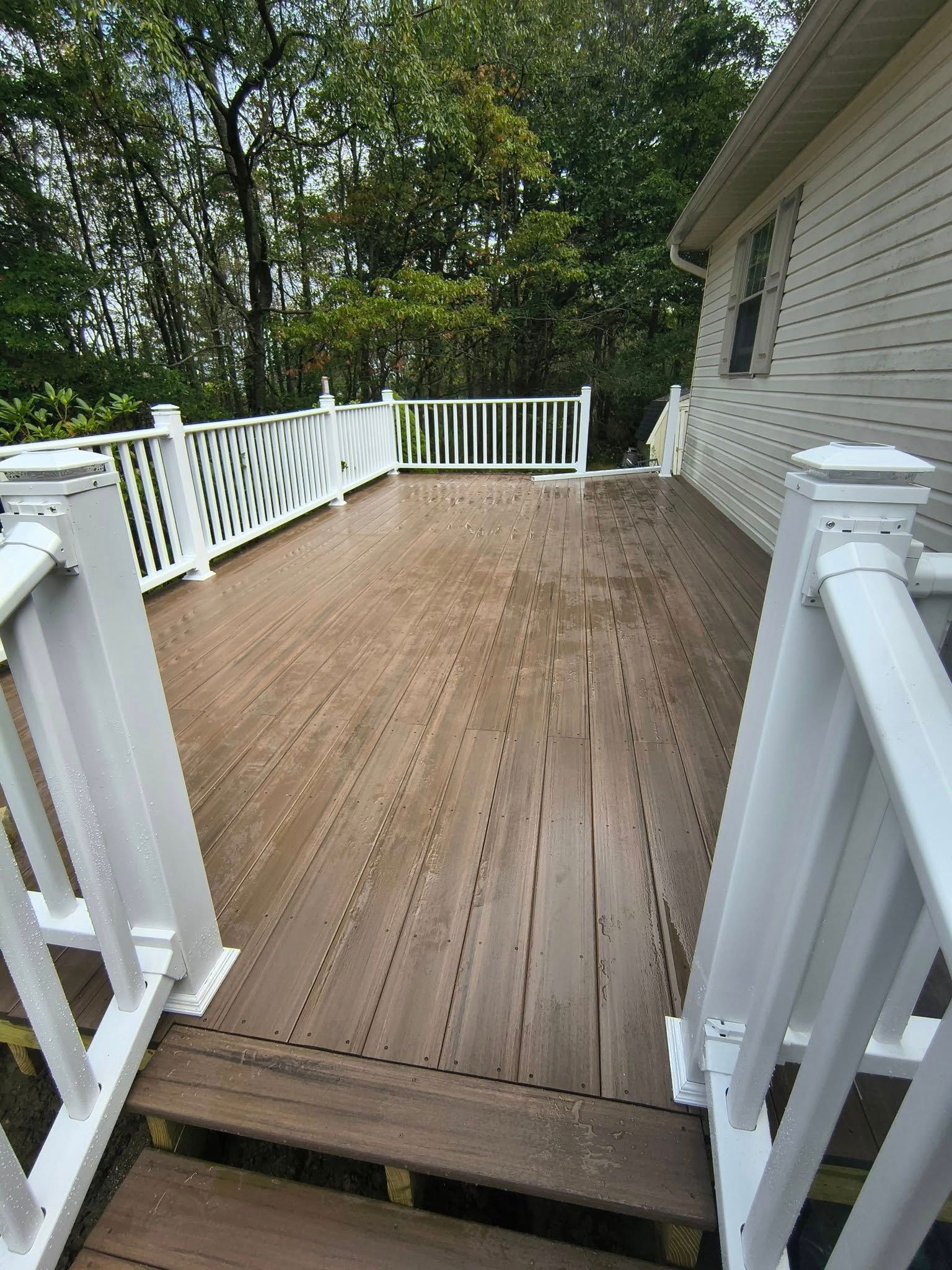 Wooden deck with white railings, attached to a light-colored house, surrounded by trees.