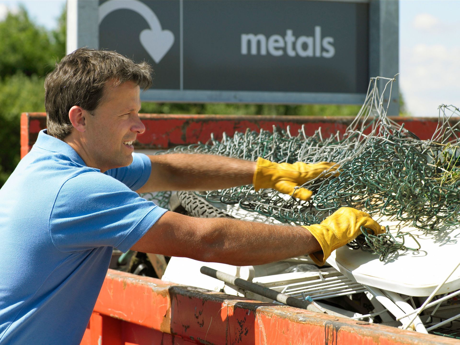 Person loading yard waste into a trailer parked on a street, with trees and houses in the background.