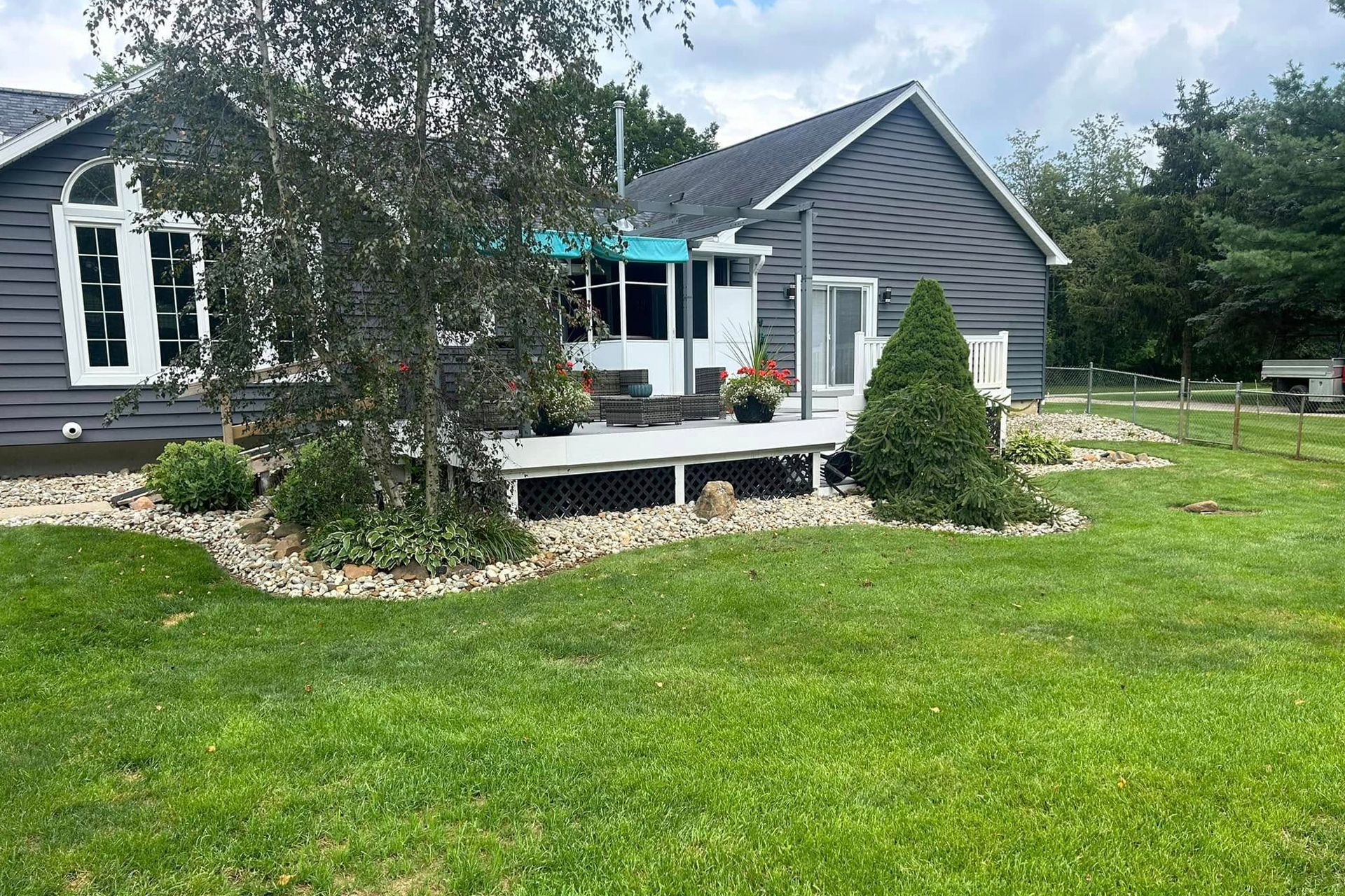 A house with a deck and garden. Gray siding, green lawn, blue awning, and flowering plants.