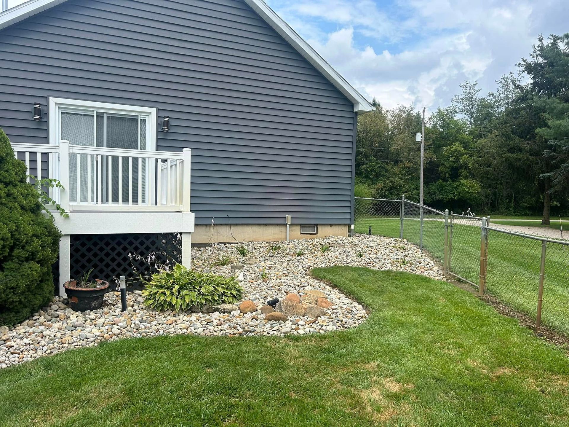 House with a deck, gravel bed, and chain-link fence in a grassy yard, under a cloudy sky.