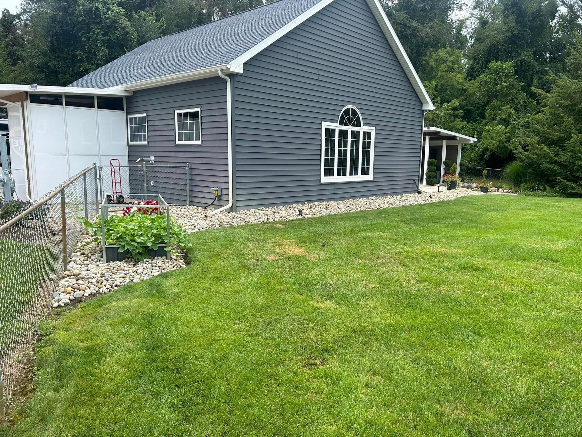 Side view of a gray house with white trim, set in a grassy yard with landscaping and chain link fence.