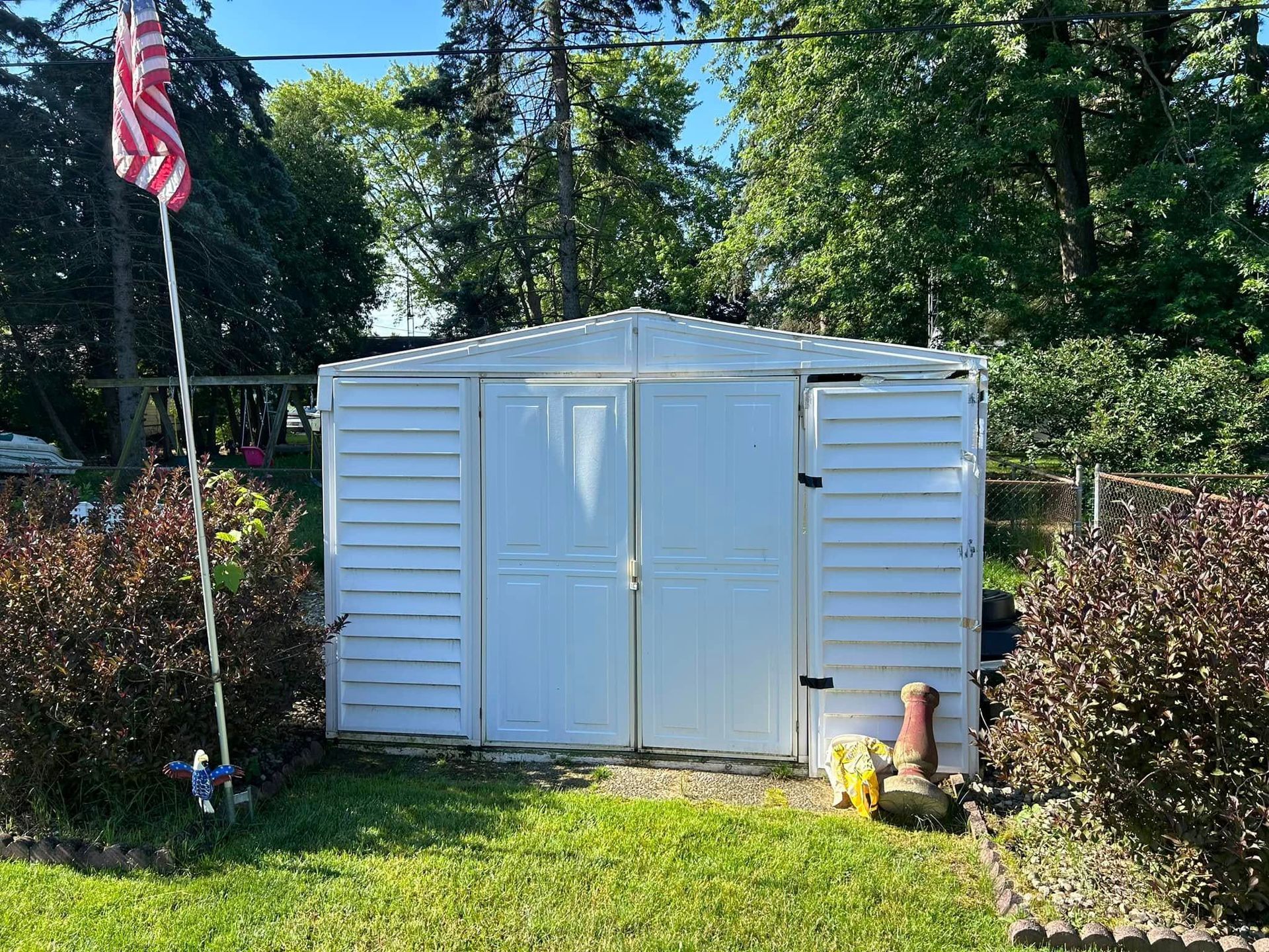 White shed with double doors, American flag, and bushes in a backyard setting.
