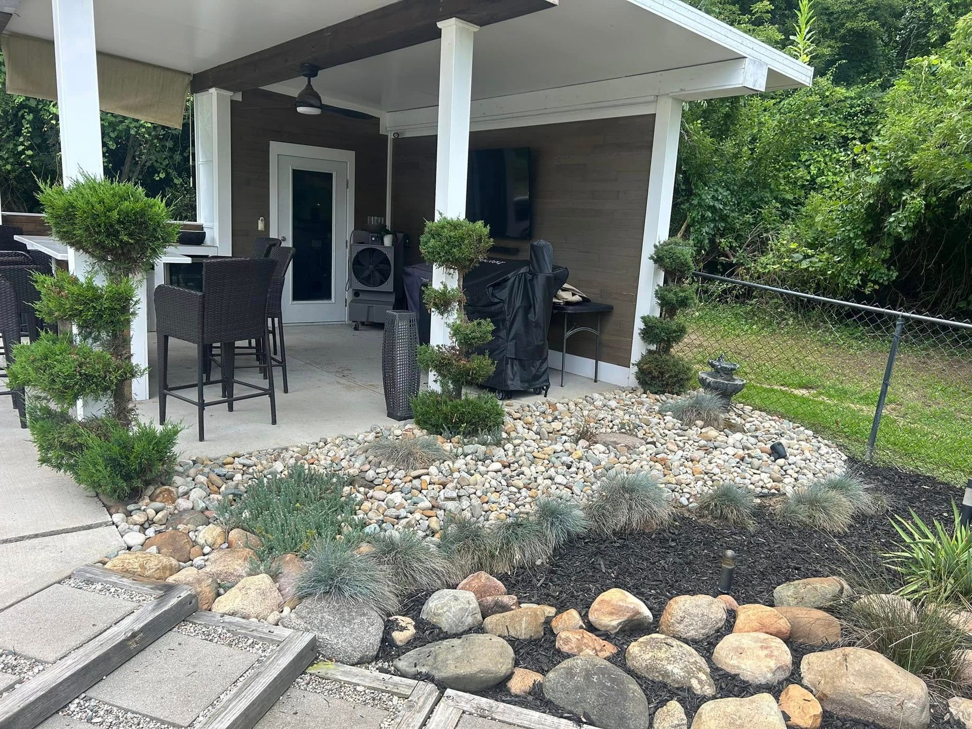 Outdoor patio with seating, a television, and small garden with rocks and shrubs.
