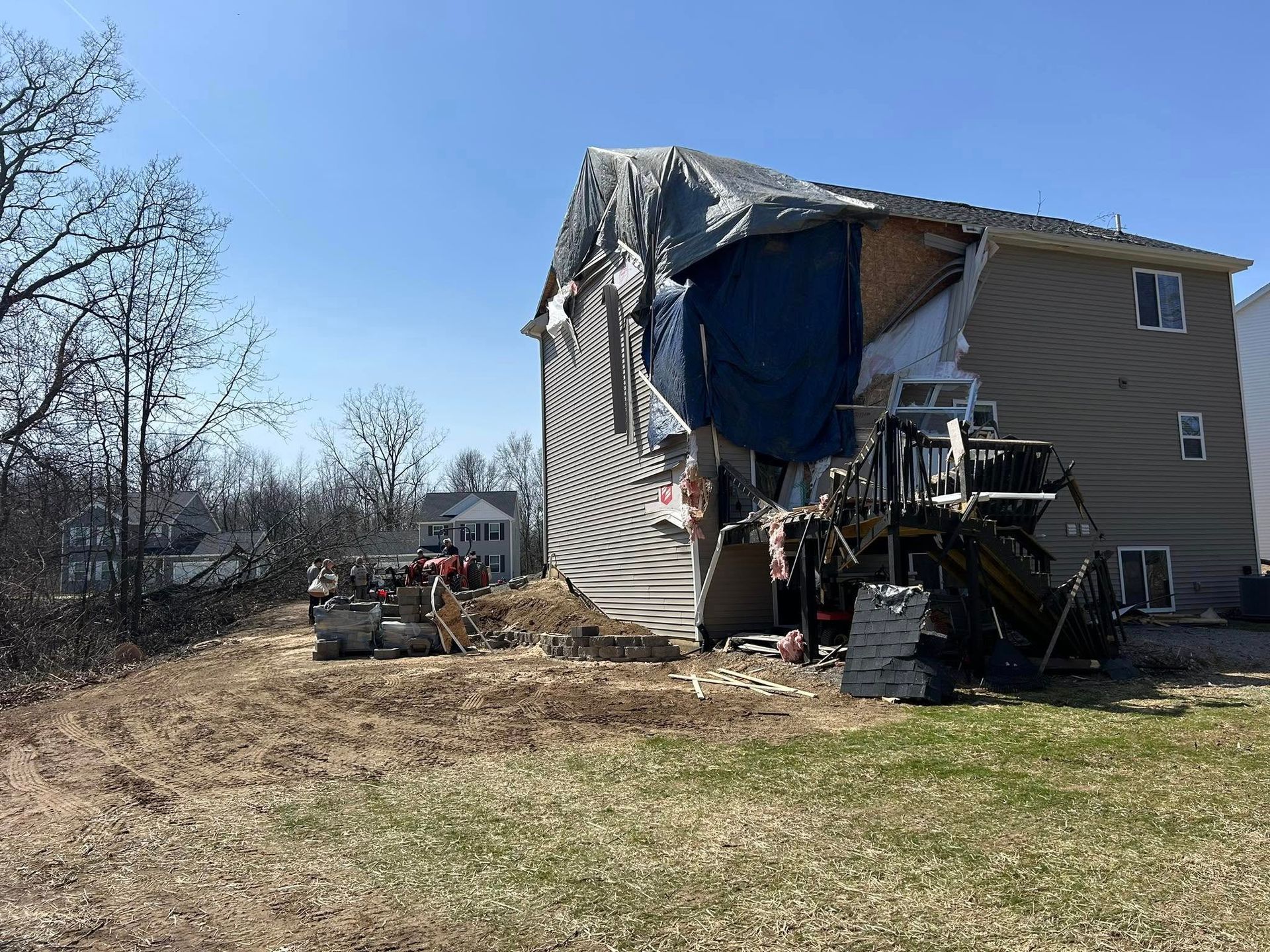 House with severe damage, missing siding and roof covered in tarp. Debris and construction equipment in yard.