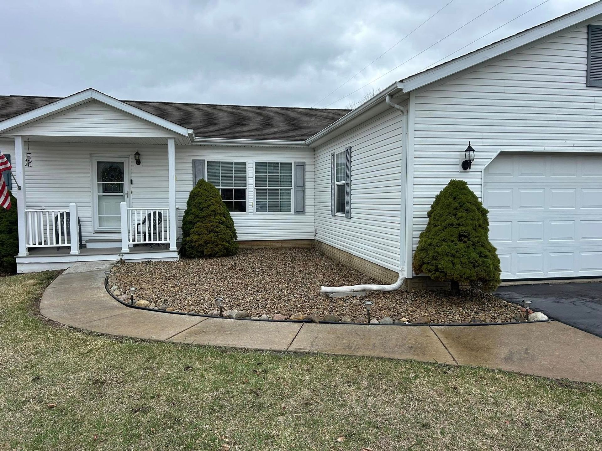 White house with a lawn and rock bed. Two evergreen bushes frame the window and garage. Cloudy sky.