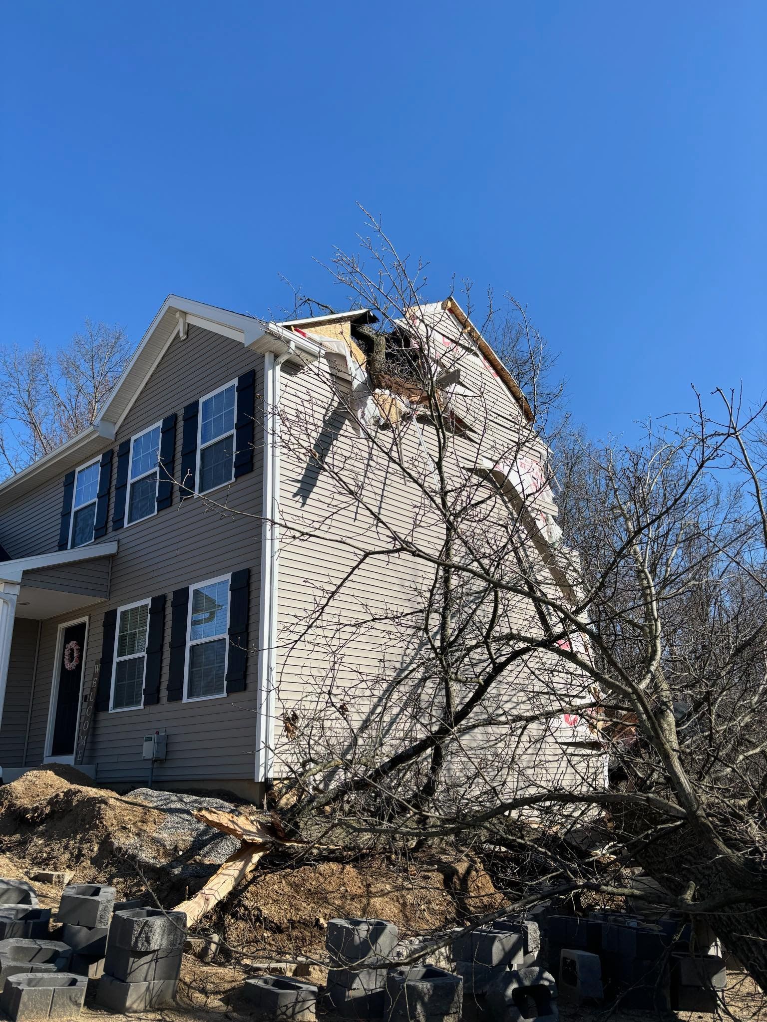 House with severe damage to the roof and side, likely from a storm. Clear sky, debris visible.
