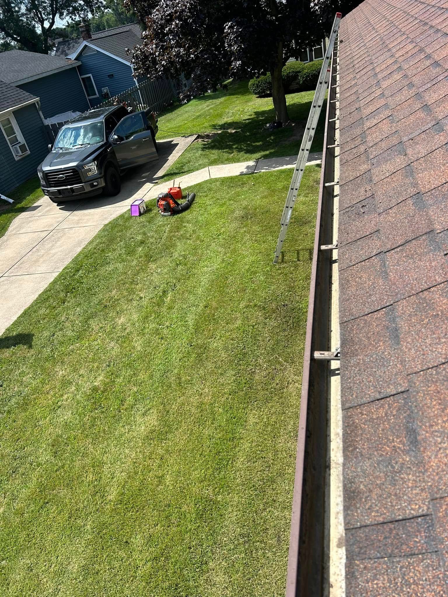 Ladder on roof gutter; person cleaning, black SUV parked in driveway. Green lawn, houses visible.