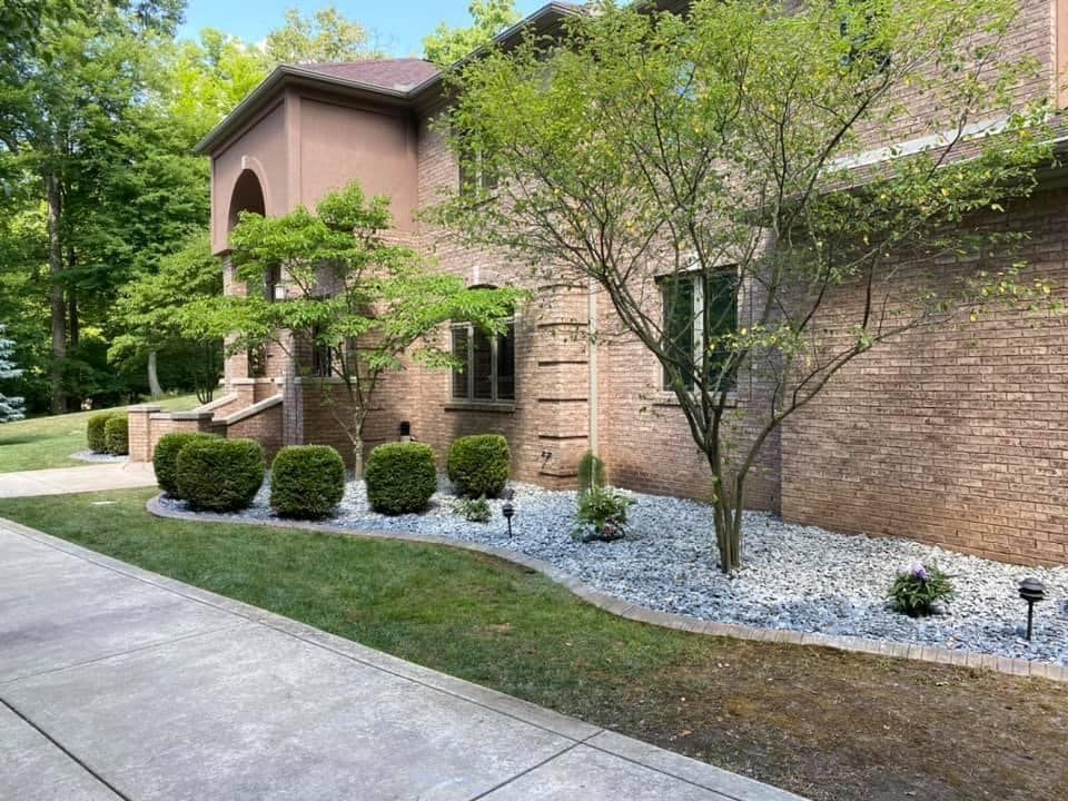 A house with a brick facade, landscaped with shrubs, trees, and light gray gravel.