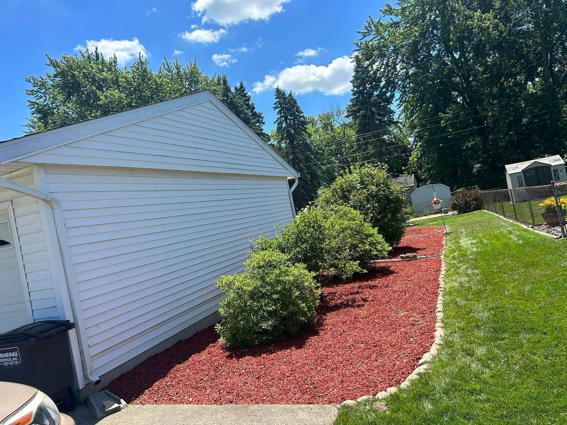 Red mulch bed with green bushes next to a white garage and green lawn on a sunny day.