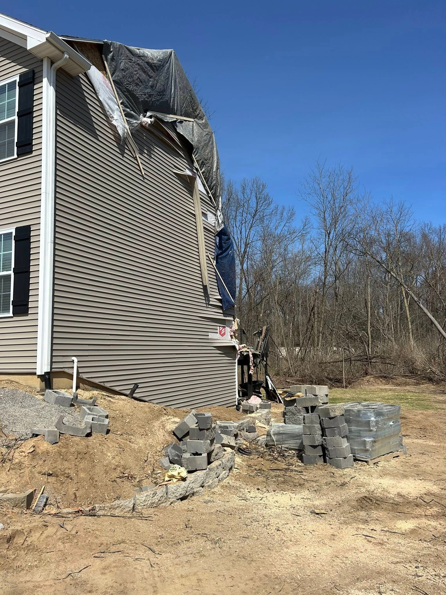 House with damaged siding and debris on dirt ground, blue sky.