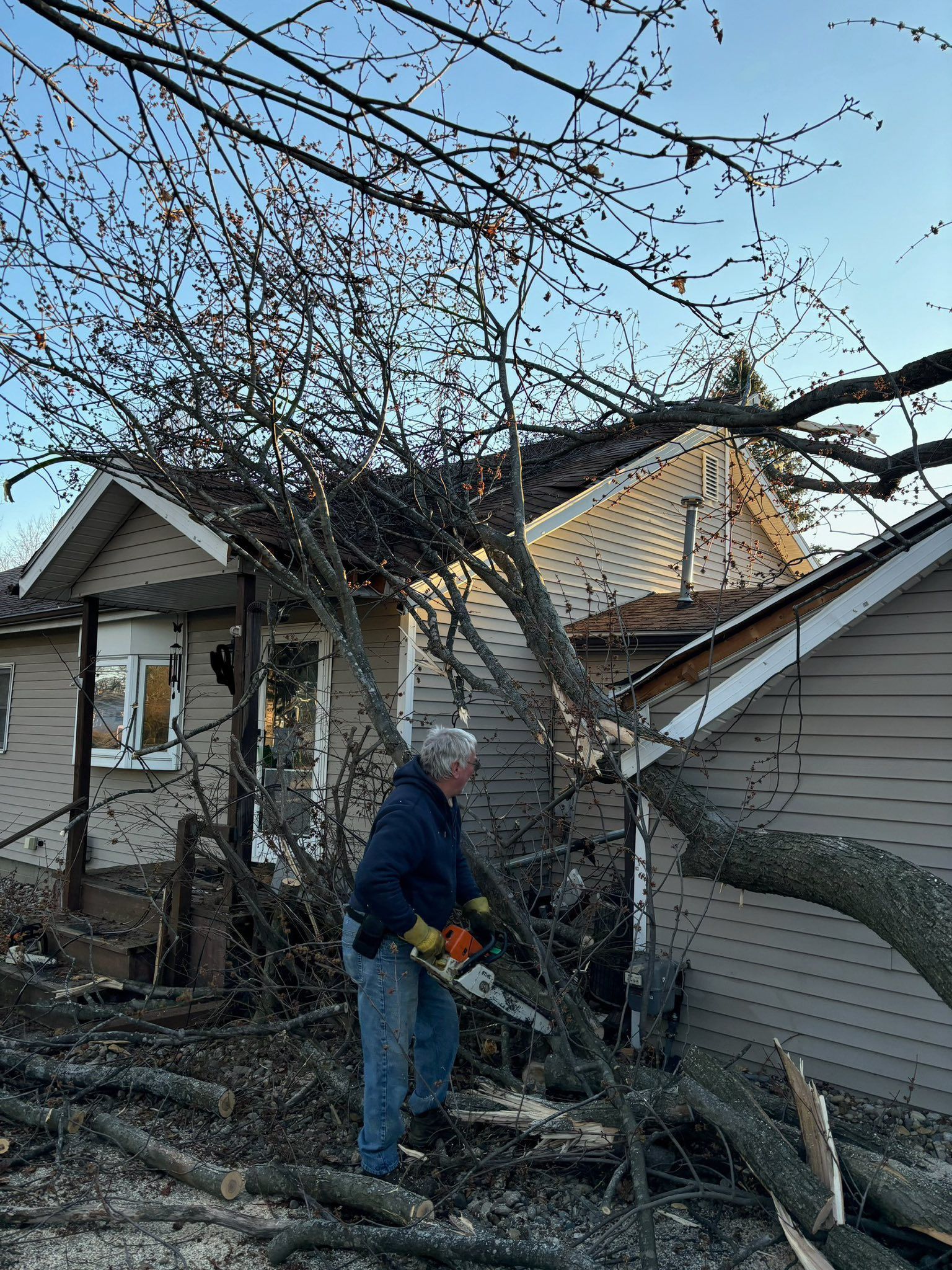 Man cuts fallen tree with chainsaw next to house. Debris and branches cover yard.