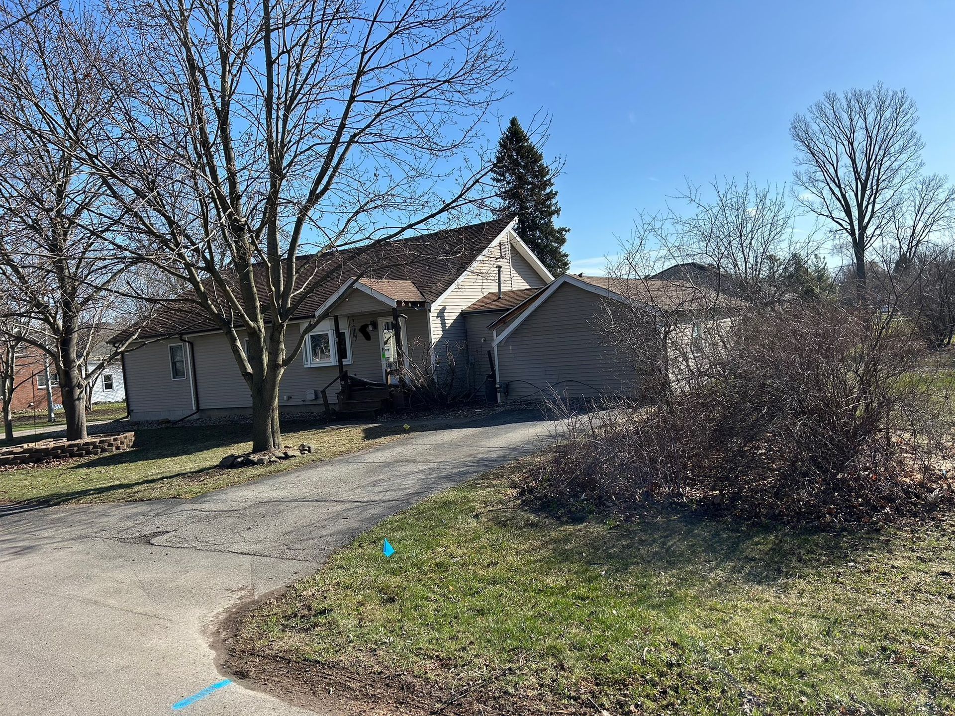 A weathered, single-story house with a driveway on a sunny day; trees partially bare.