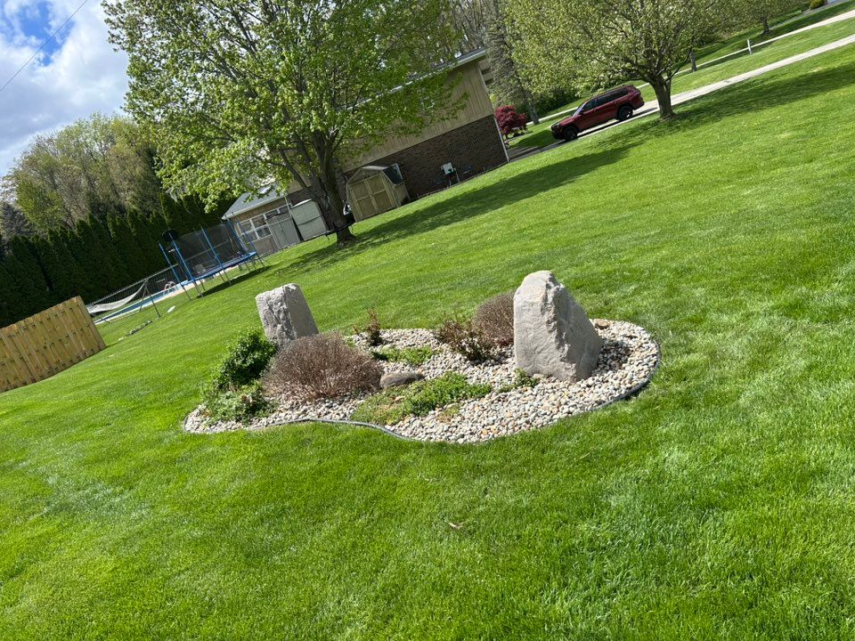 Grassy yard with a small rock garden featuring two upright stones, surrounded by light-colored rocks and plants.