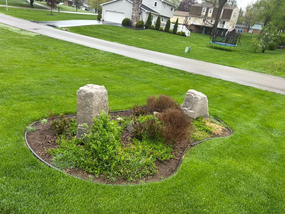 Stone markers in a small garden bed on a grassy lawn, with a paved path and houses in the background.